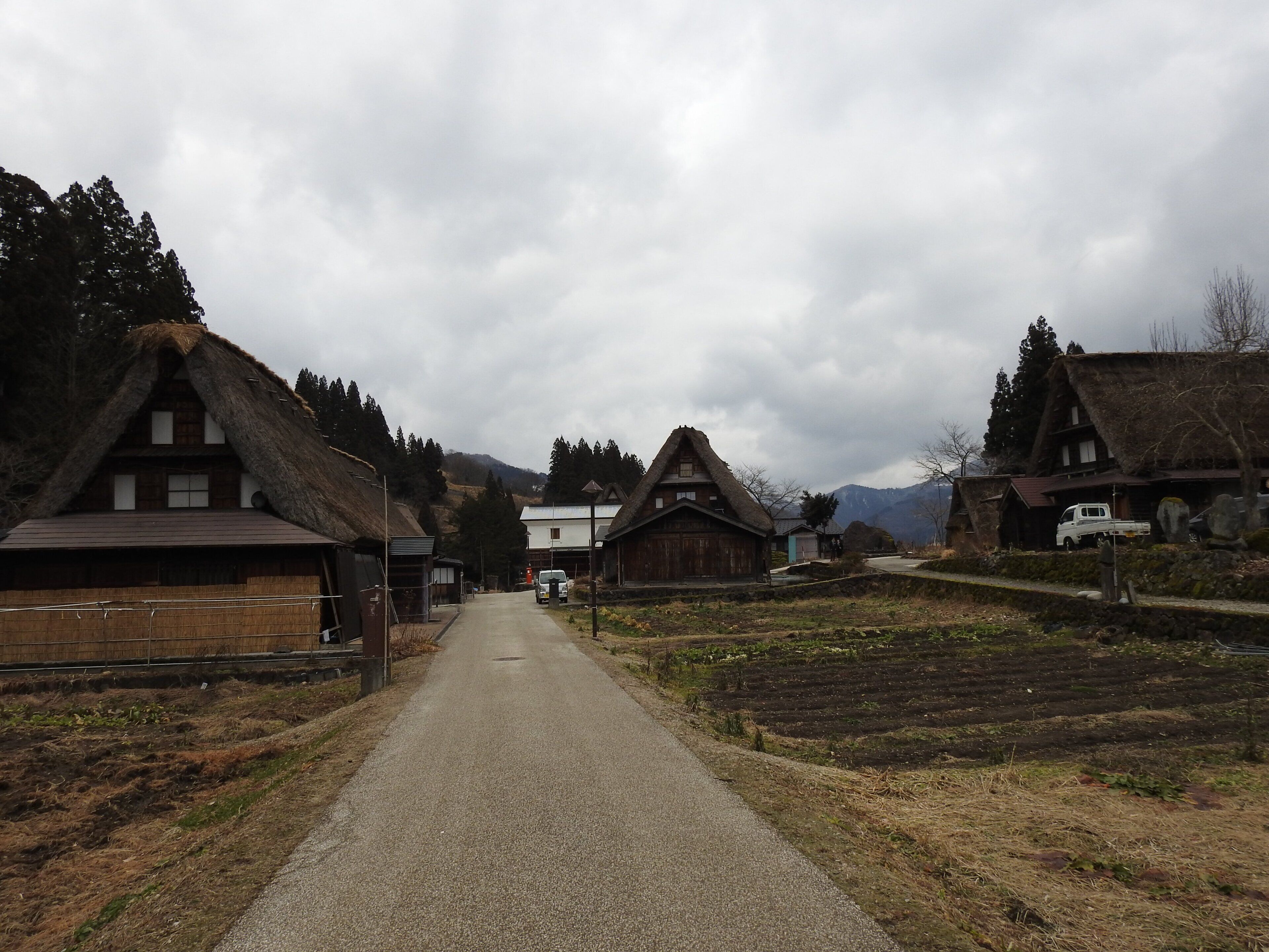 A rural village with traditional thatched-roof houses, a paved road, and a mountainous backdrop.