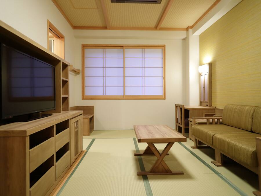 Una habitación de estilo japonés tradicional con suelo de tatami, un televisor sobre un soporte de madera, una mesita baja, un sofá y una puerta corrediza con ventanal.