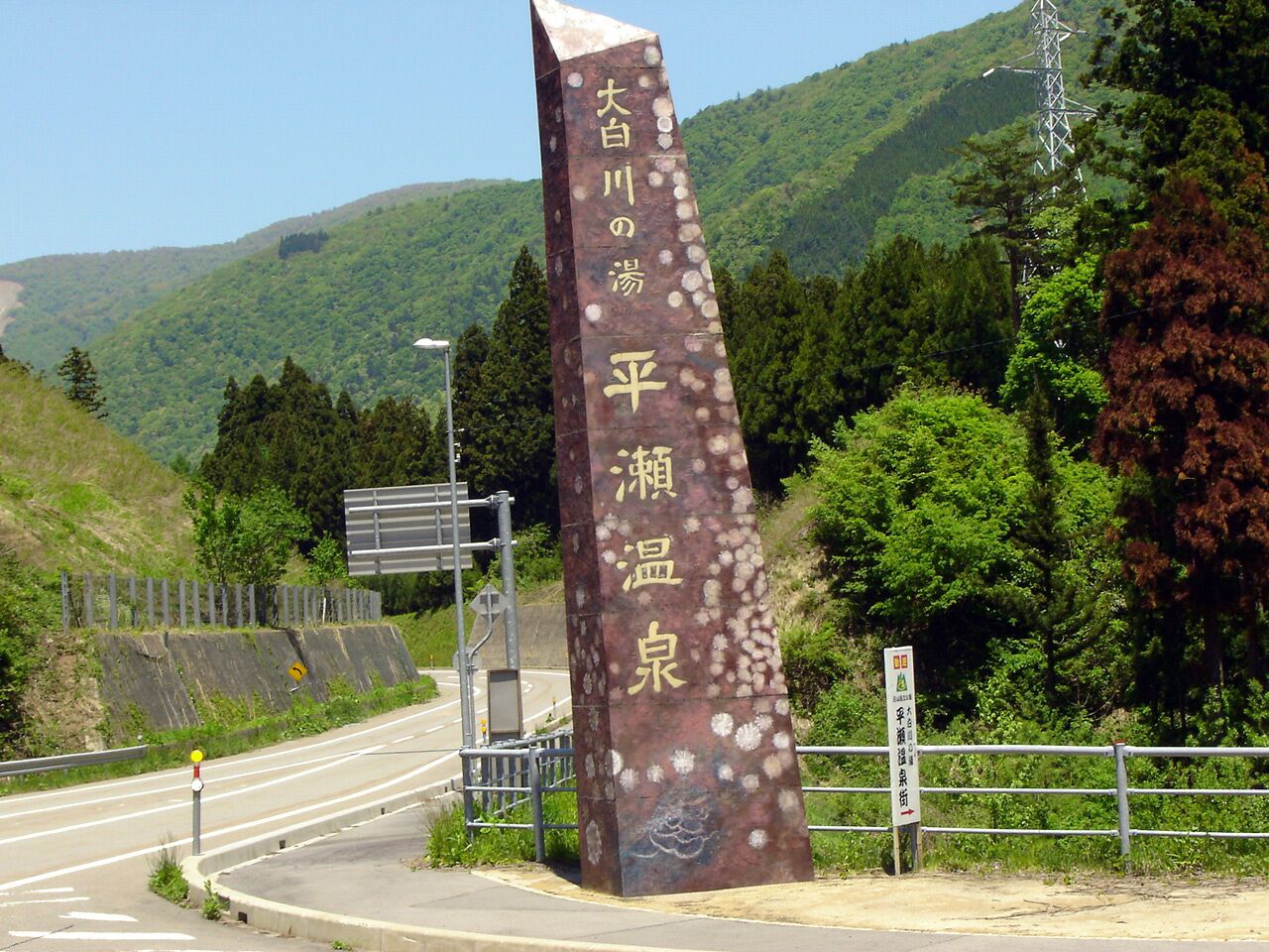 Un cartel alto y vertical con caracteres japoneses, situado junto a una carretera con barandilla y un paisaje montañoso de fondo.