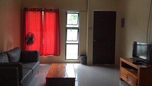 A living room with a sofa, a wooden coffee table, a television on a wooden stand, a fan, and a window with red curtains. - Cris Place Dumaguete (Valencia)
