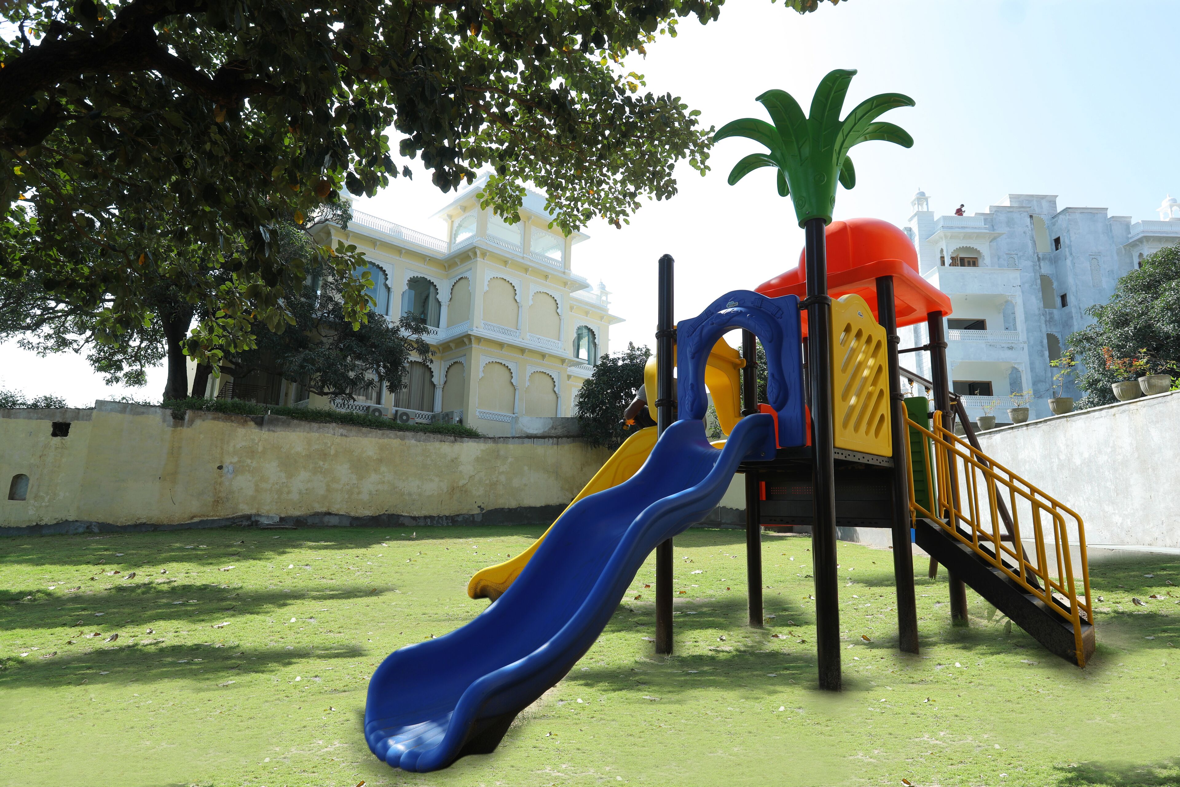 Un parque infantil con toboganes y una estructura de escalada, rodeado de césped y un edificio con balcones al fondo.