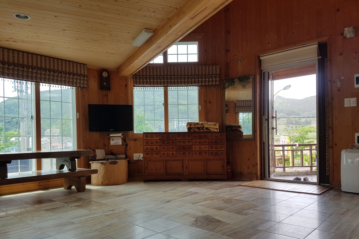 A spacious room with wooden walls and a large window, a flat-screen TV, a wooden cabinet, and a bench.