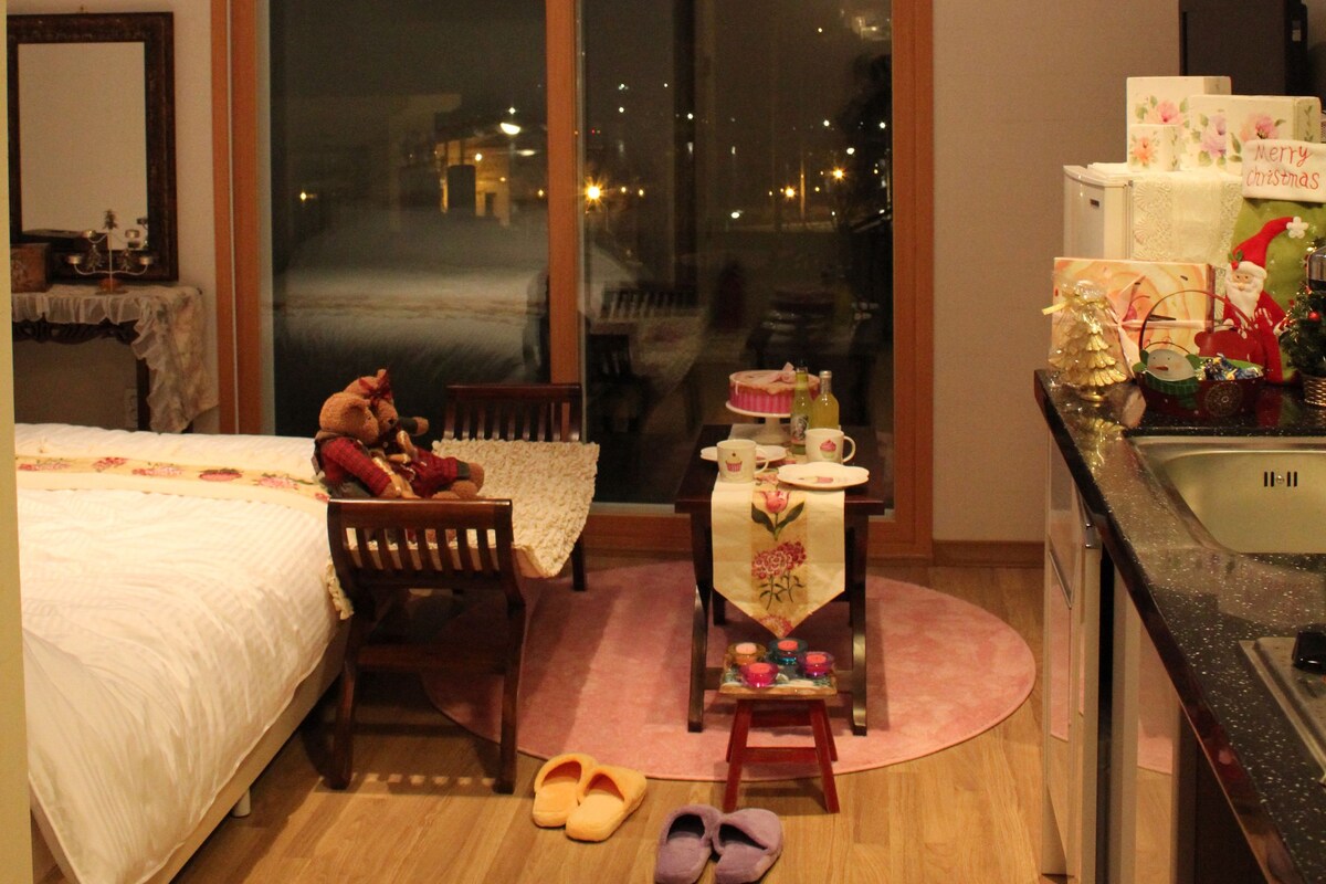 A bedroom with a bed, a chair, a small table with a tablecloth, a pair of slippers, and a view of the city through a large window.