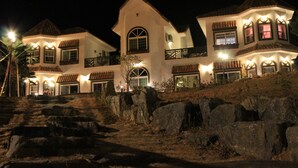A large, multi-story building with a white facade and multiple balconies, situated on a hillside with large rocks in the foreground. - Red Praha Pension (Geoje)