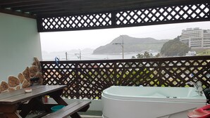 A balcony with a wooden table, a green bucket, a white bathtub, and a view of a mountain and buildings. - Red Praha Pension (Geoje)