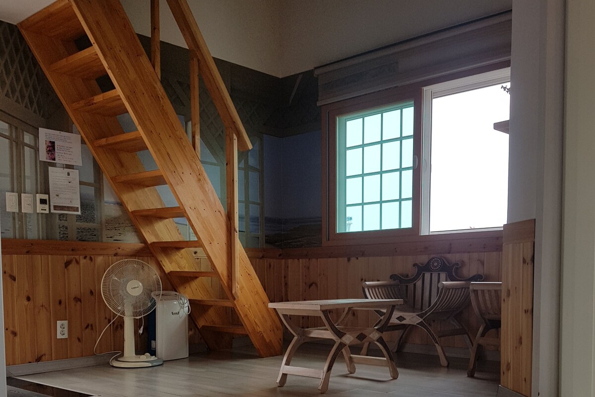 A room with a wooden staircase, a dining table with chairs, a fan, and a window with a view.