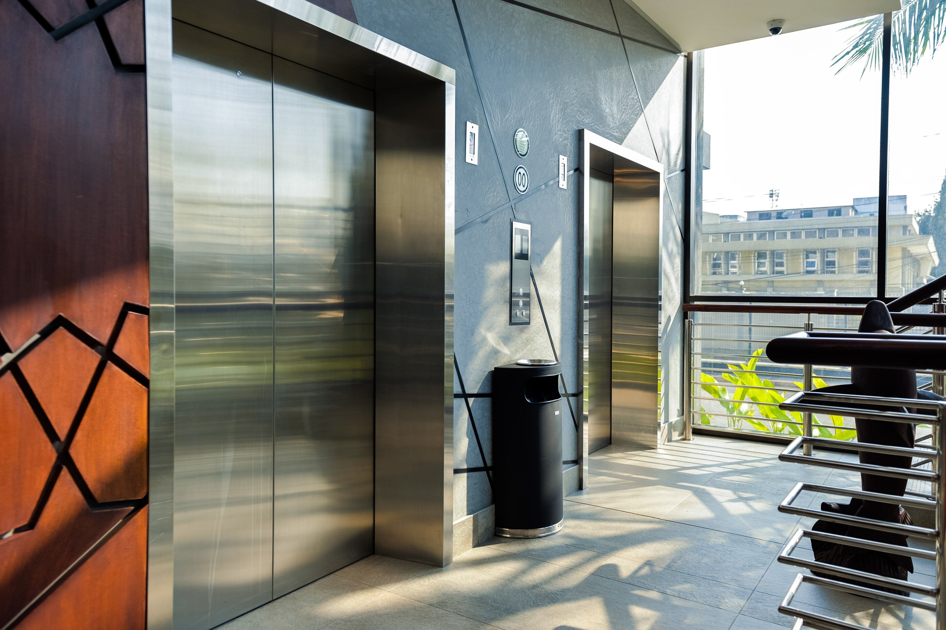 A modern elevator with metallic doors, a trash bin, and a staircase leading to a balcony with a view of buildings.
