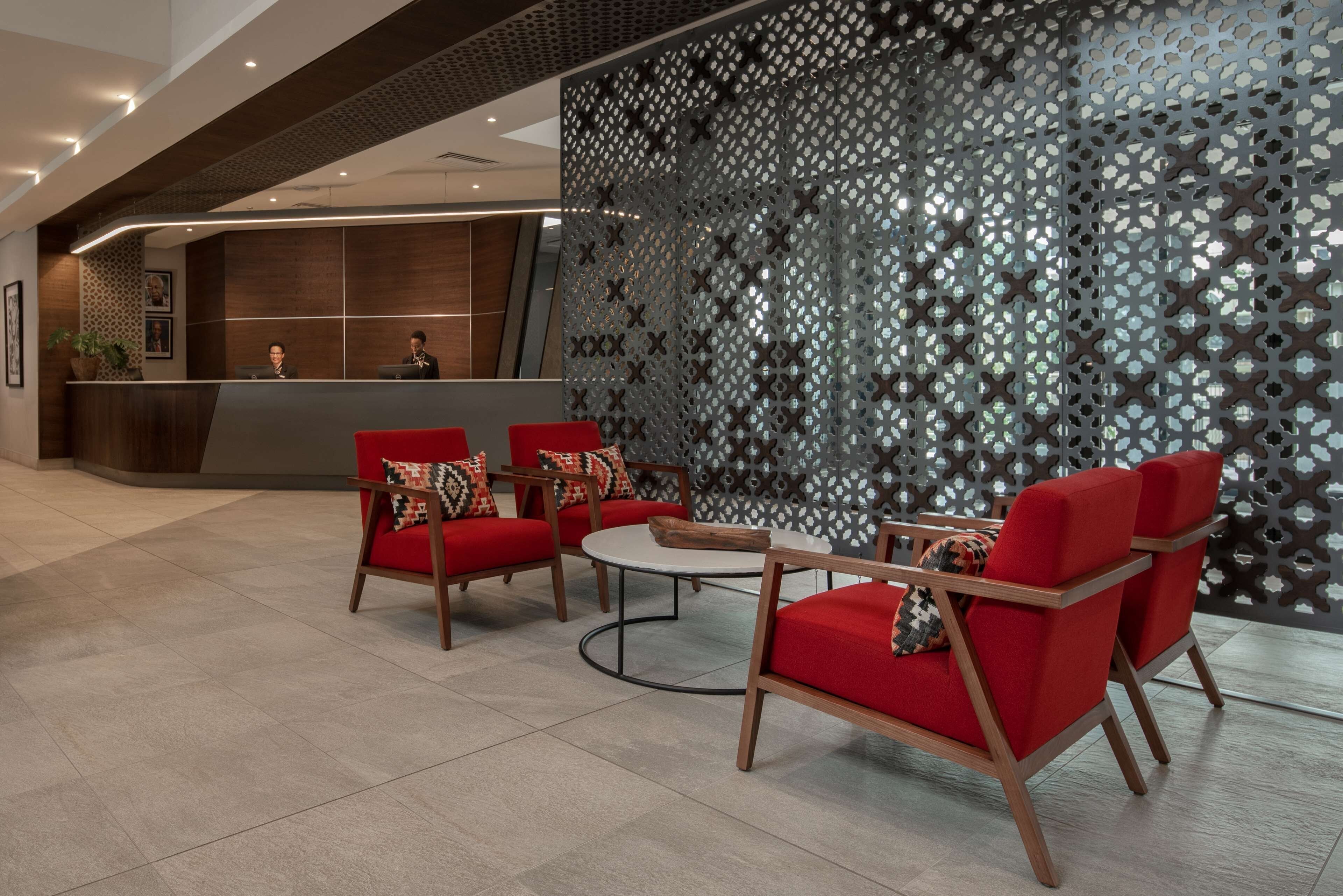 A modern reception area with a reception desk, red upholstered chairs, a round coffee table, and a decorative wall with a geometric pattern.