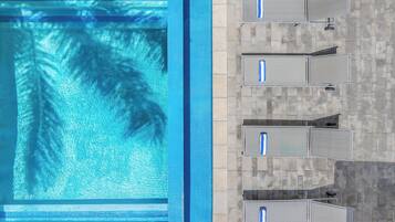 An aerial view of a swimming pool with clear water and palm tree reflections, adjacent to a tiled patio area with multiple lounge chairs.