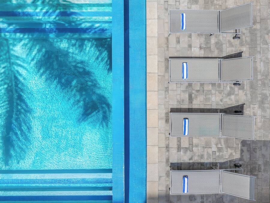 An aerial view of a swimming pool with clear water and palm tree reflections, adjacent to a tiled patio area with multiple lounge chairs.