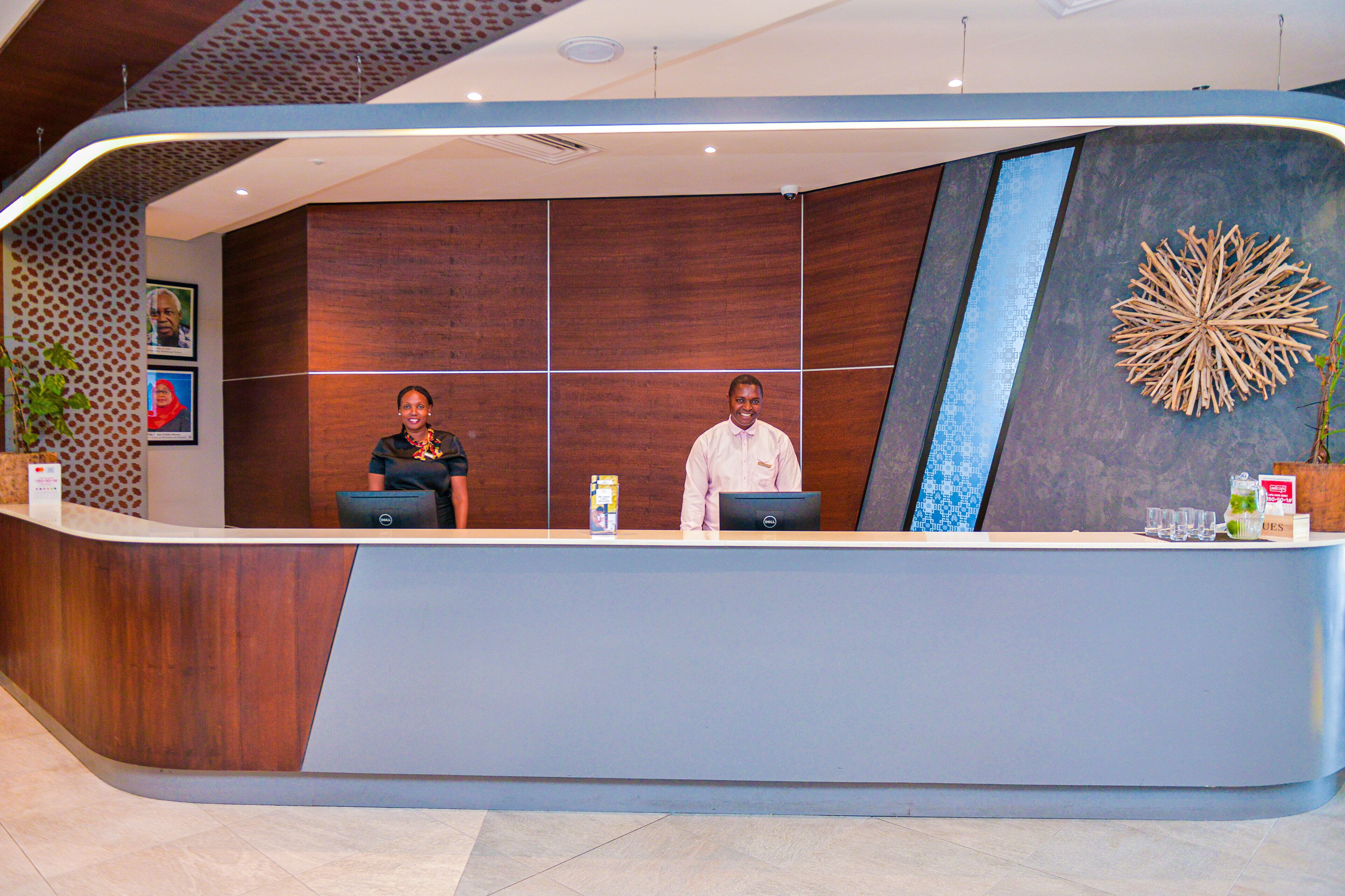 A reception desk with two staff members, a wooden panel wall, and a decorative wall piece.