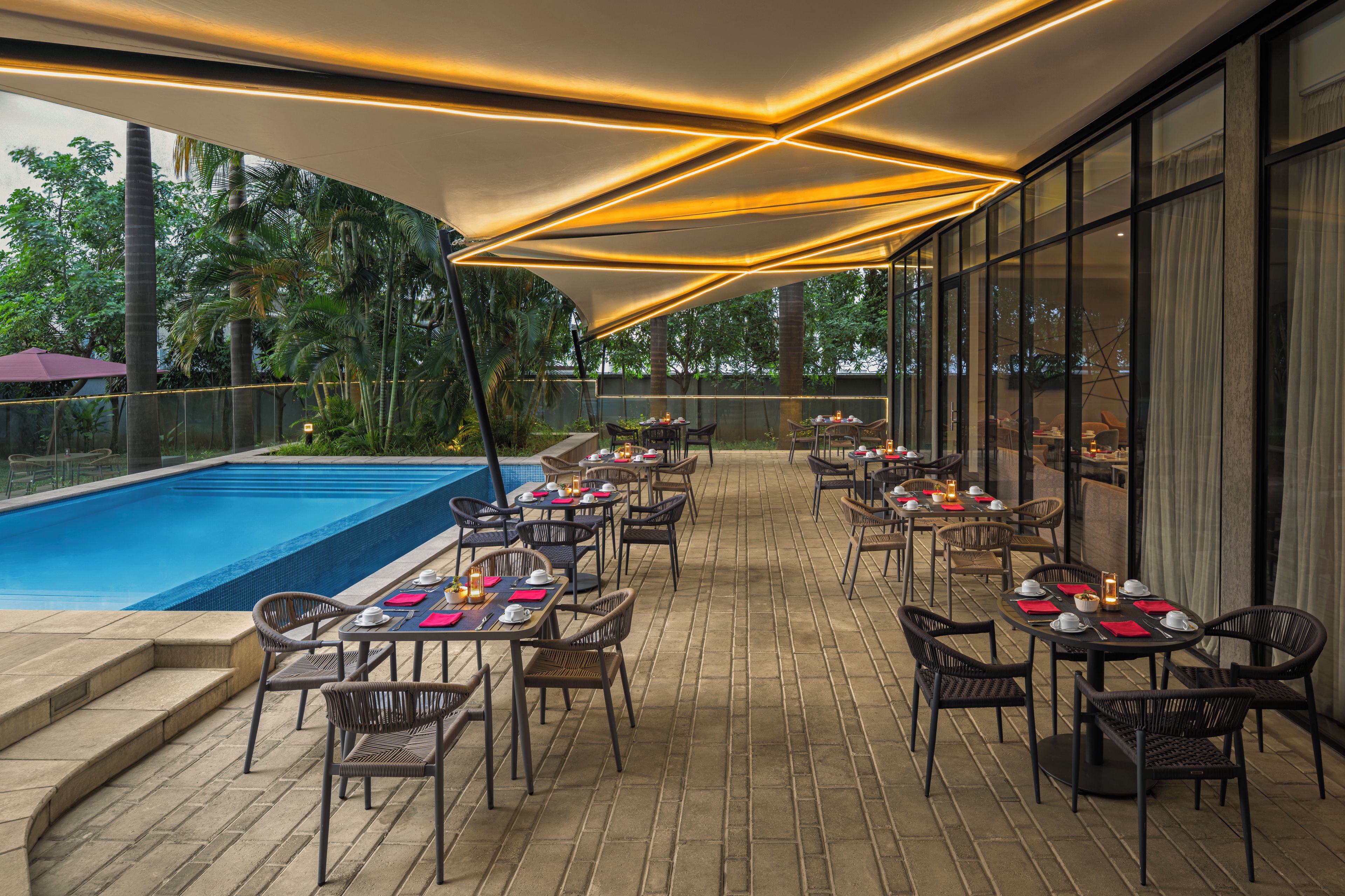 An outdoor dining area with tables and chairs, a swimming pool, and a view of greenery.