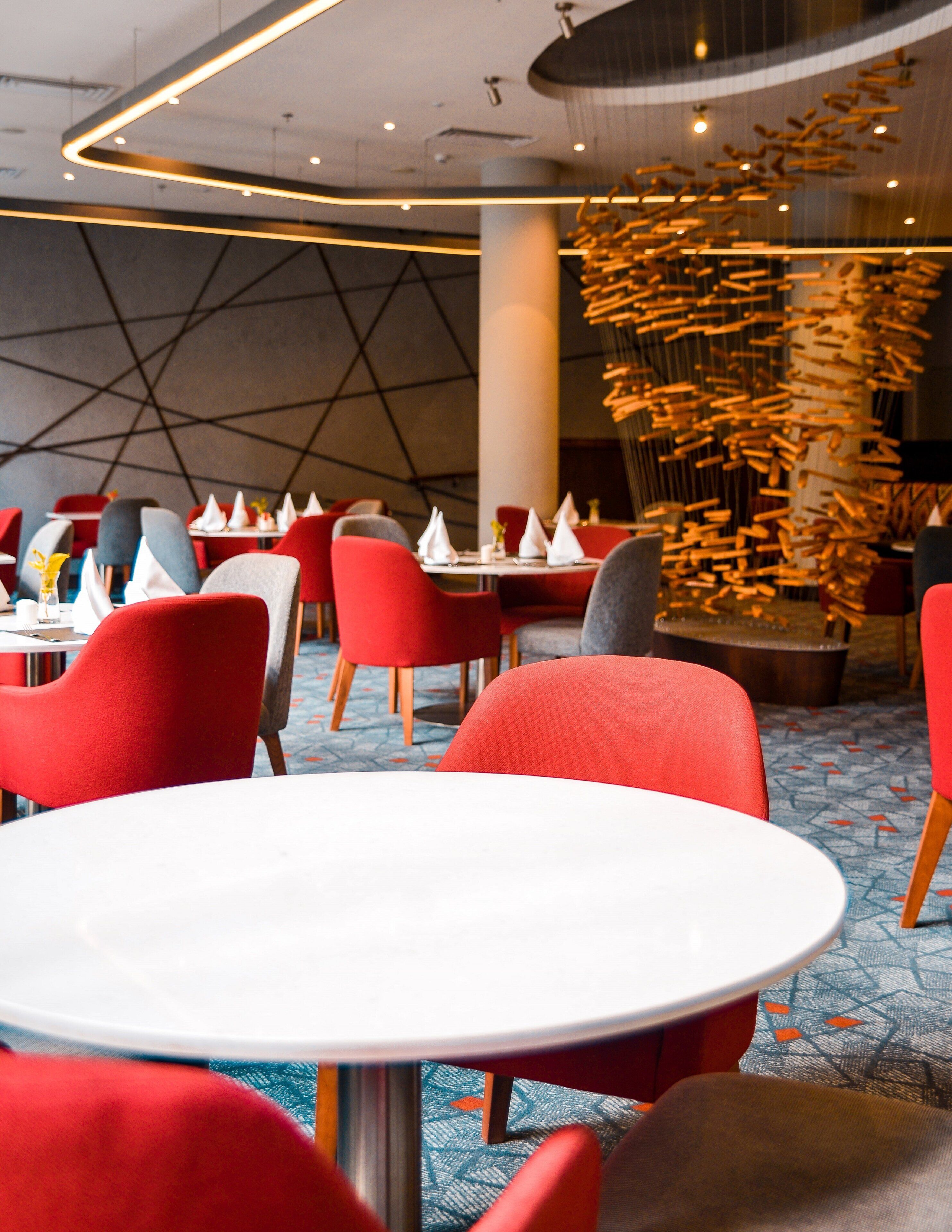 A modern dining area with red chairs and white tables, featuring a unique ceiling light fixture and geometric wall design.