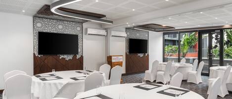 Un salón de conferencias con mesas redondas, sillas blancas, un televisor de pantalla plana y un cartel que dice "Crowne Plaza".