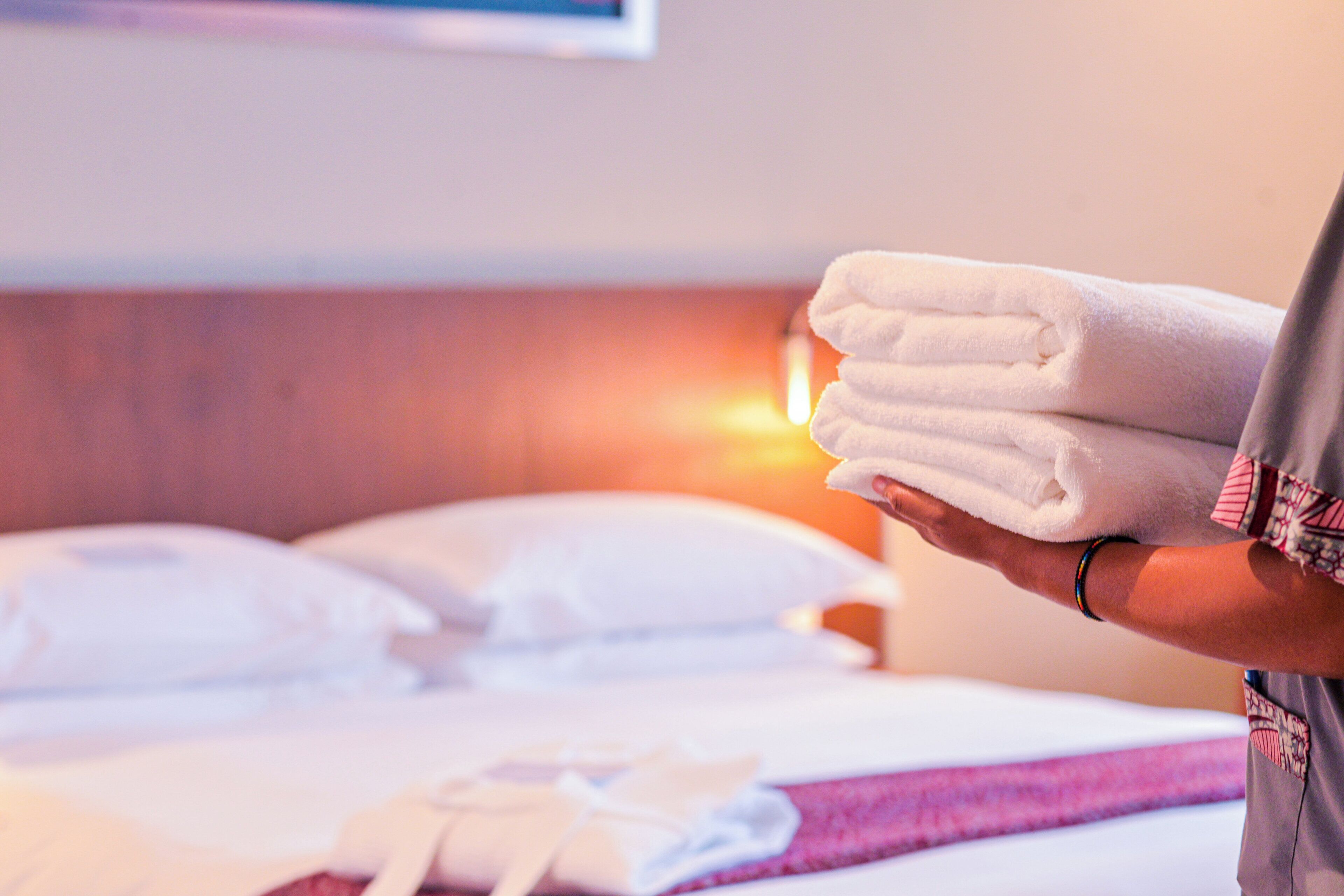 Una mano sosteniendo una pila de toallas dobladas, una cama con sábanas blancas y un cabecero con una fuente de luz.