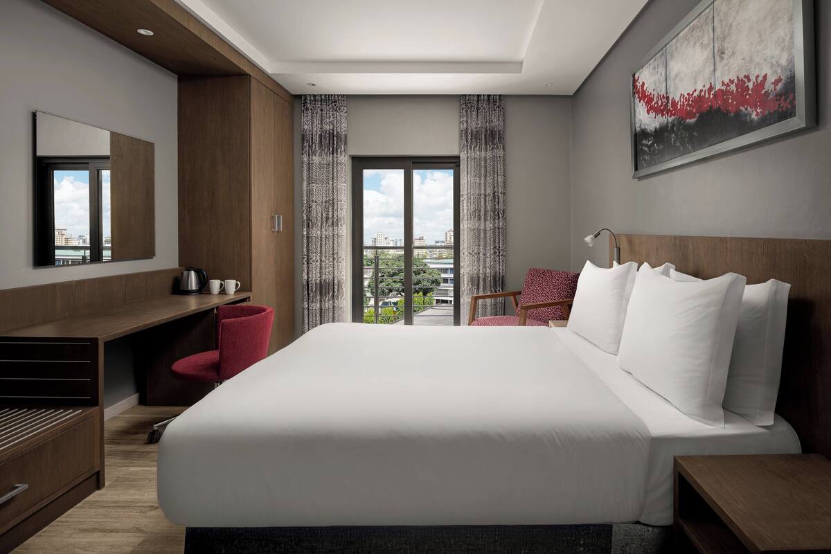 Une chambre d’hôtel moderne, dotée d’un grand lit, d’un bureau, d’une chaise et offrant une vue sur la ville par la fenêtre.