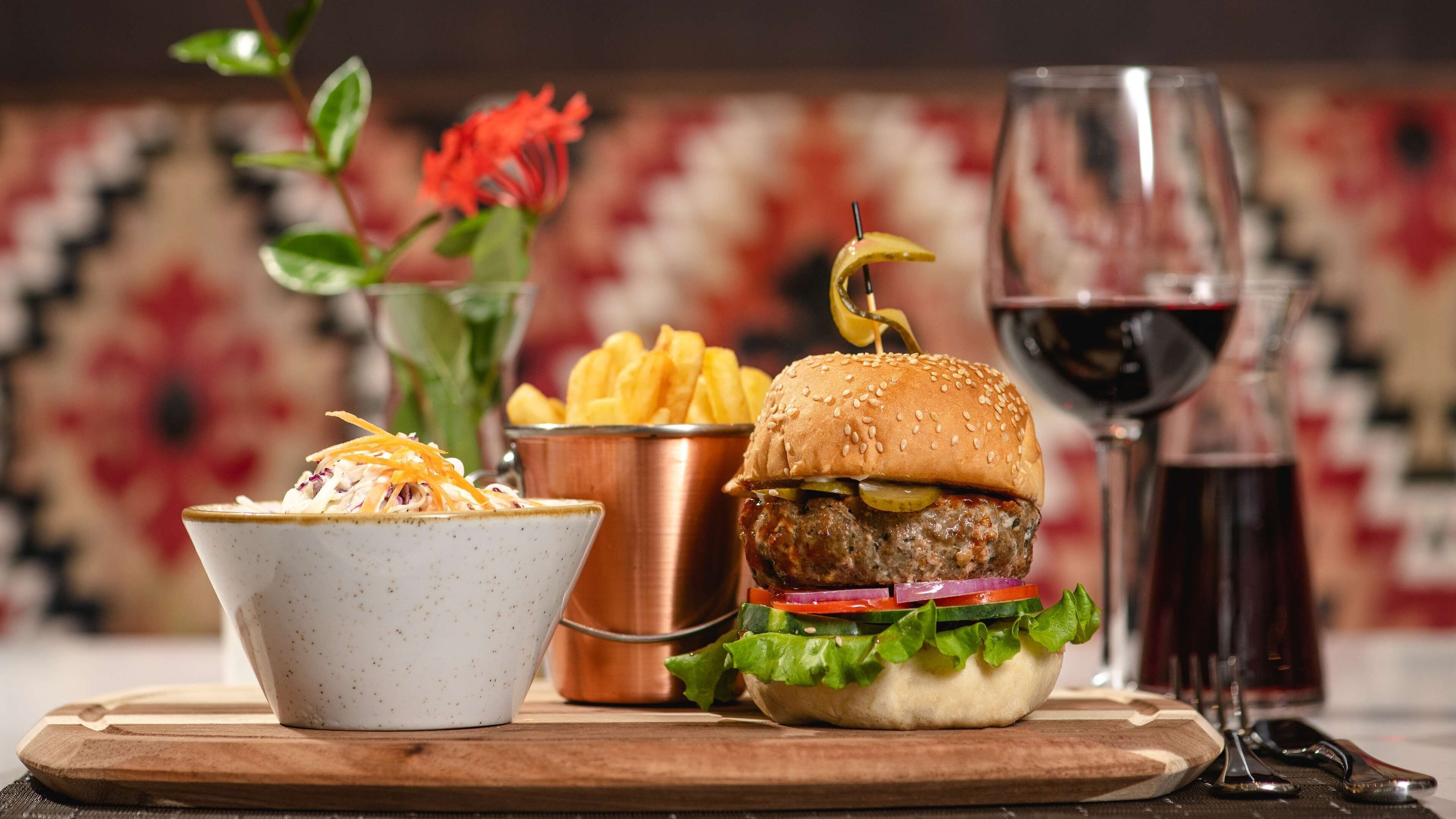A burger, fries, and a salad on a wooden board with a glass of red wine and a copper pot in the background.