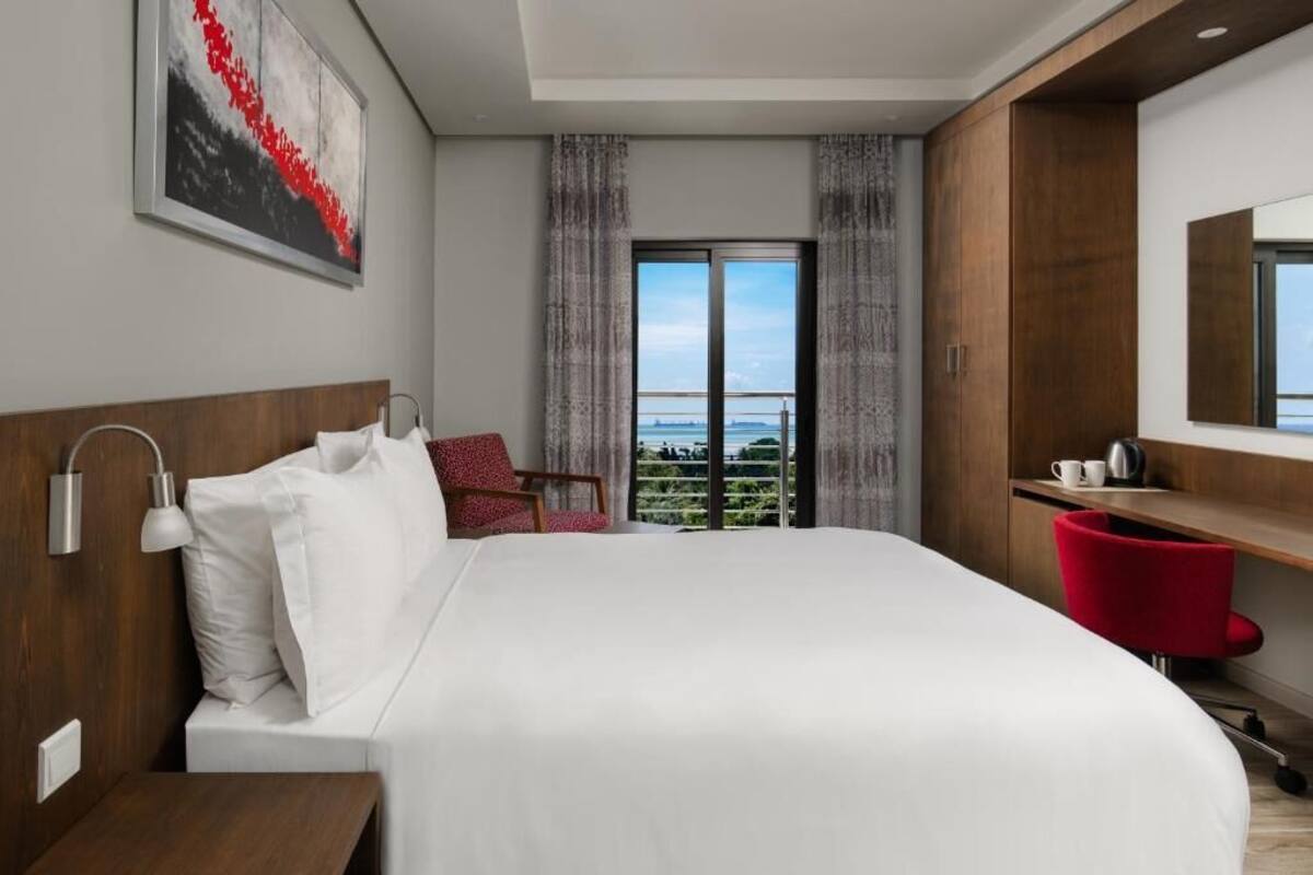 A modern hotel room with a large bed, a desk, a chair, a mirror, and a view of the outdoors.
