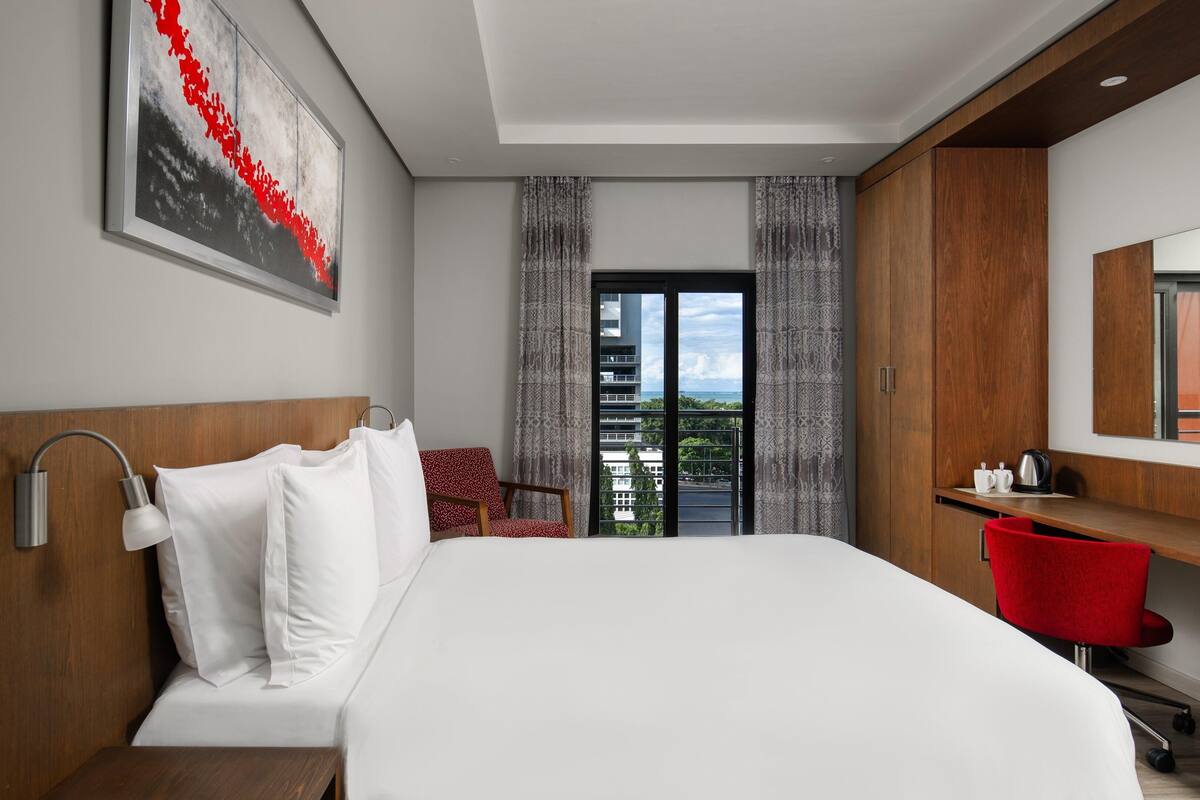 A modern hotel room with a bed, a desk, a chair, a large window, and a painting on the wall.