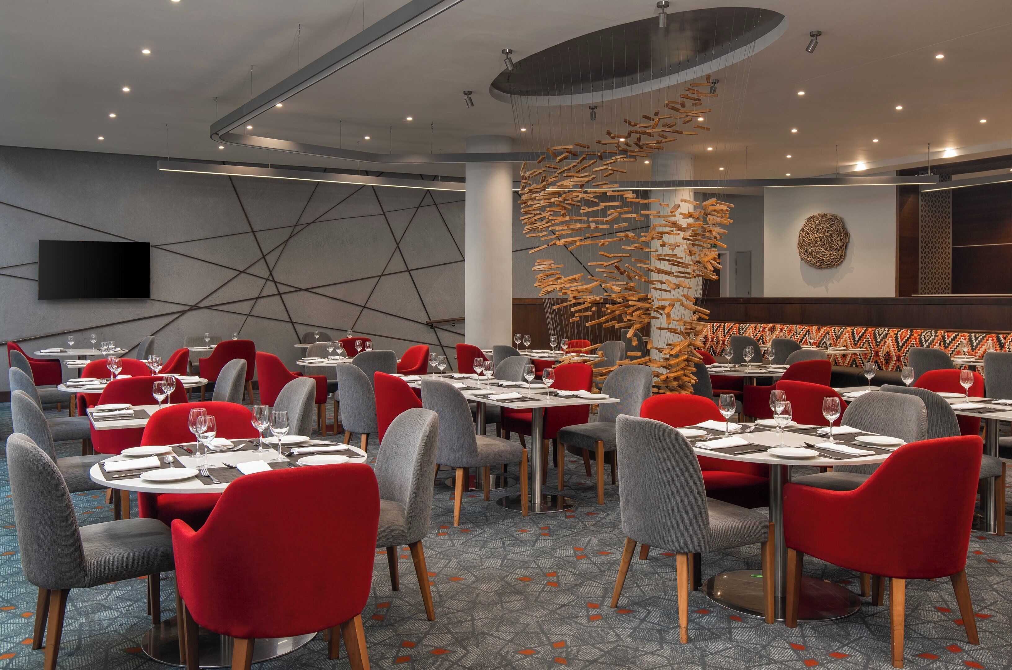 Un restaurante moderno con mesas redondas, sillas rojas y grises, y una original instalación artística de madera.
