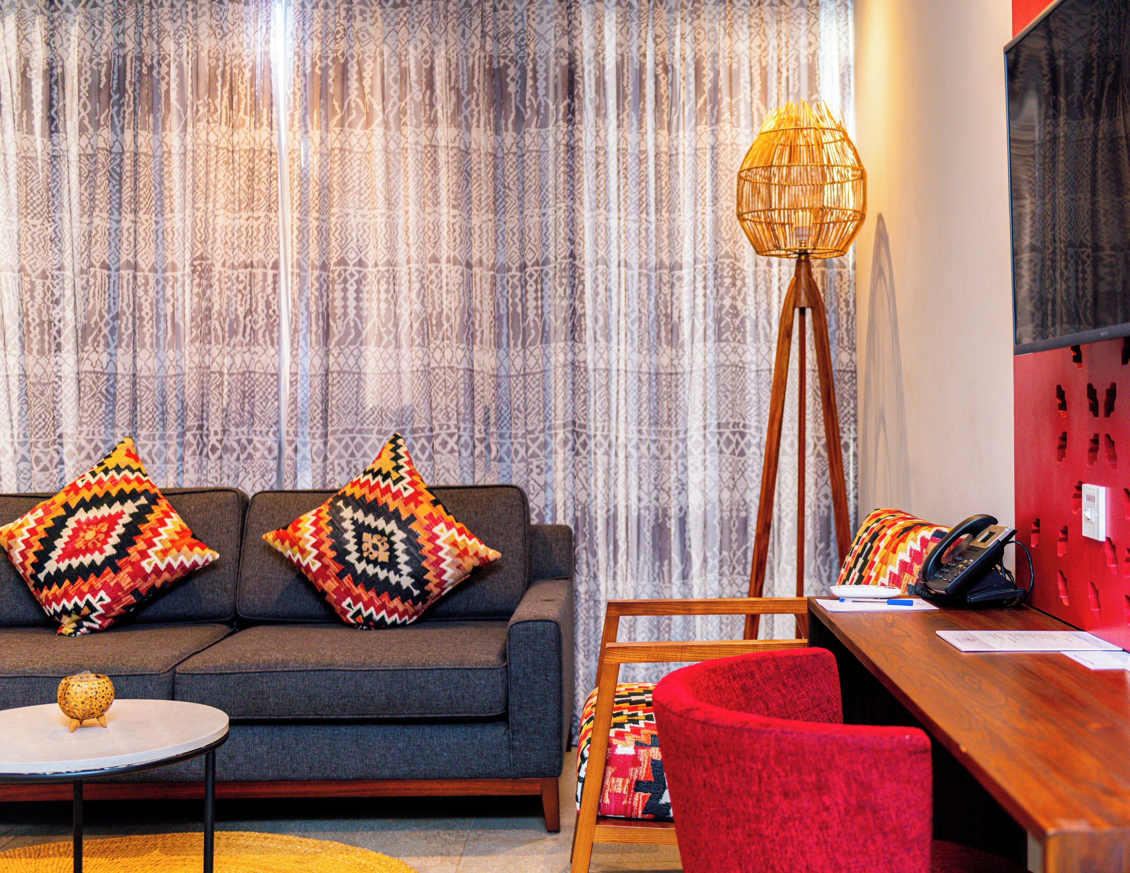 A living room with a sofa, two patterned cushions, a small table, a red chair, a floor lamp, and a wooden desk with a telephone and papers.