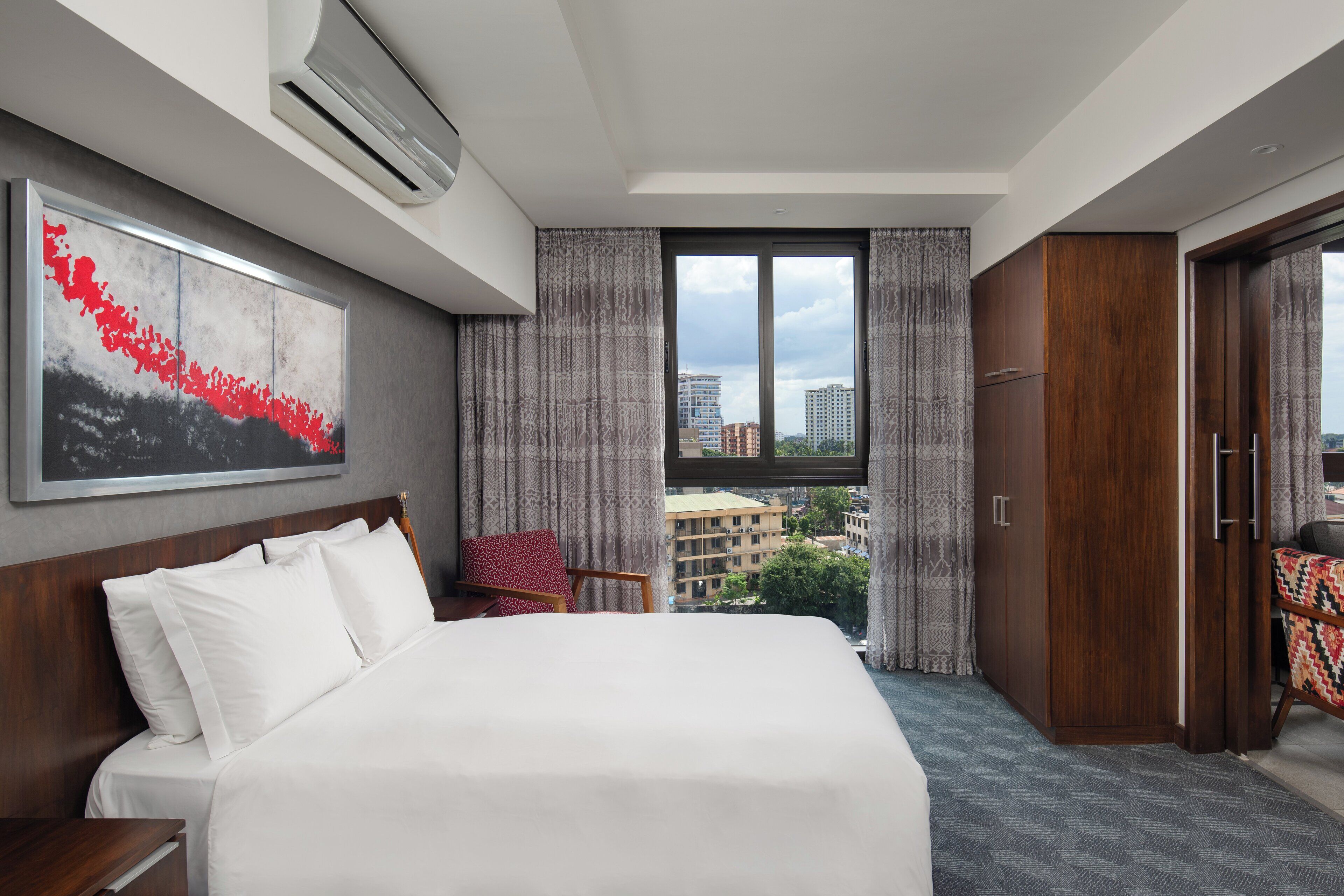 A hotel room with a large bed, a desk, a chair, a wardrobe, and a window with a city view.