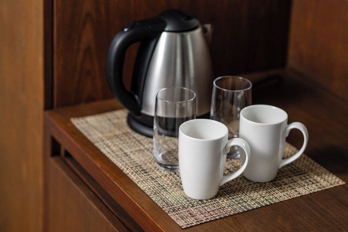 Une table en bois surmontée d’un set de table en fibres tressées, une bouilloire électrique noire et argentée, deux verres transparents et deux tasses blanches.