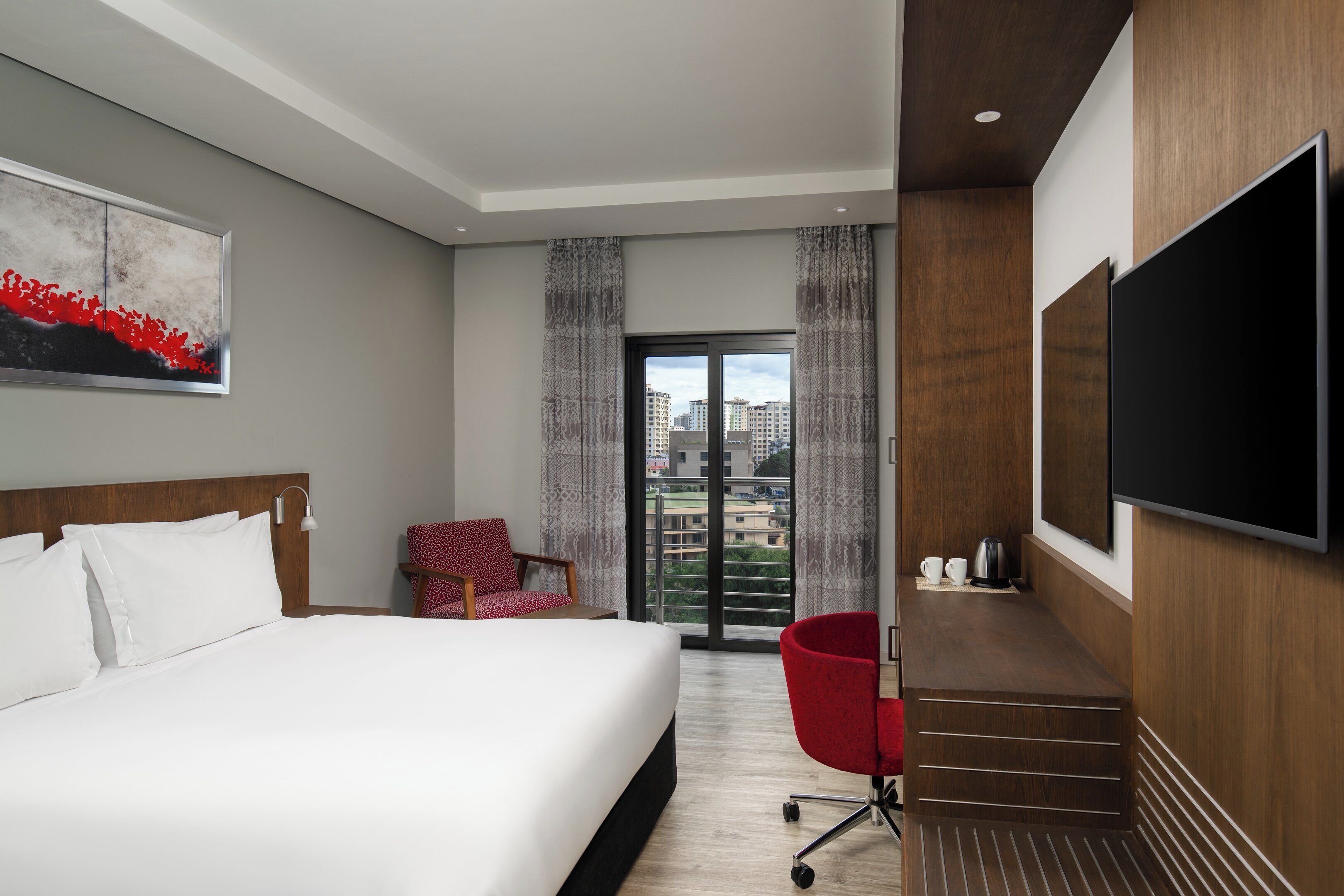 A hotel room with a bed, a red chair, a TV, and a balcony with a city view.
