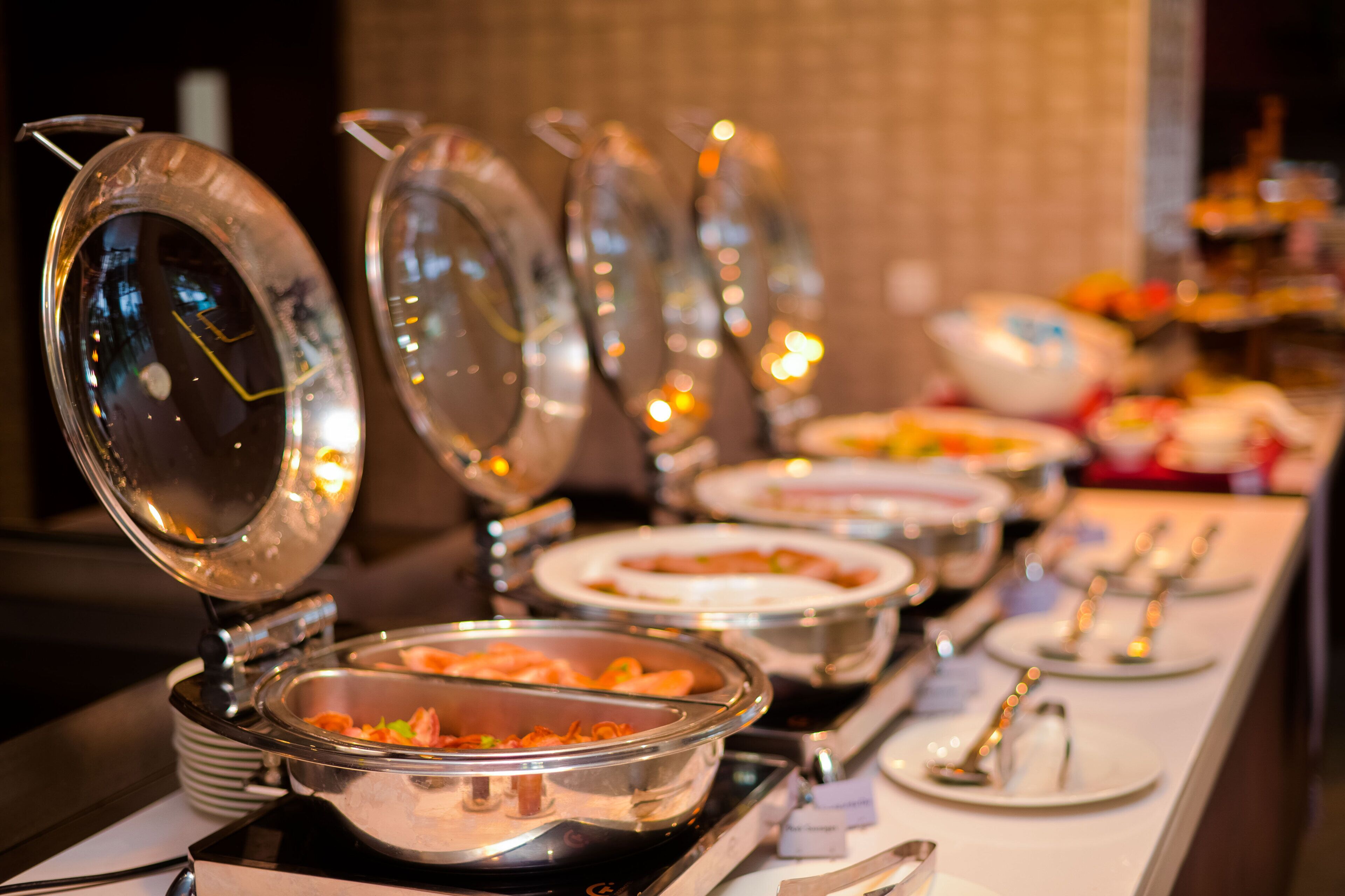 Ein reichhaltiges Buffet mit mehreren Servierstationen, an denen jeweils eine abgedeckte Speise und Besteck bereitstehen.