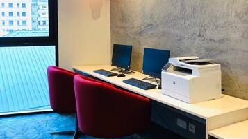 A modern office space with two desks, each with a computer monitor, keyboard, and printer. There are two red chairs and a window providing a view of buildings outside.
