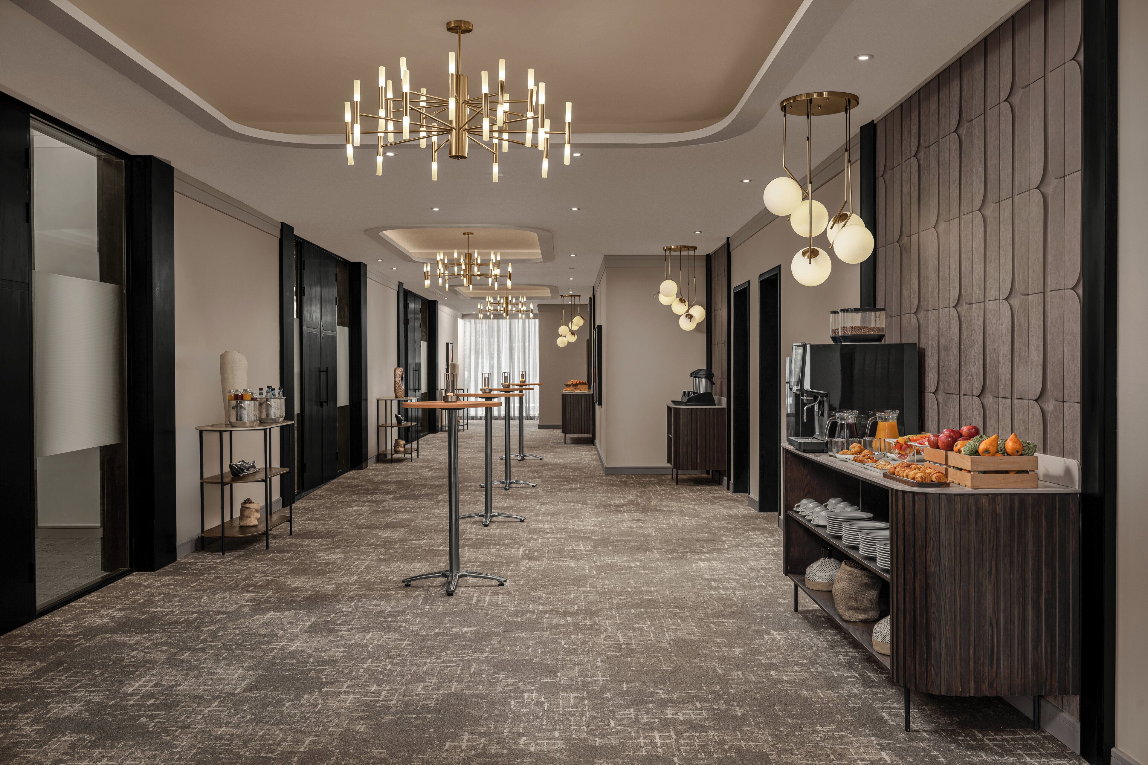 A modern hotel lobby with a central reception desk, multiple seating areas, and a buffet with fruits and beverages.