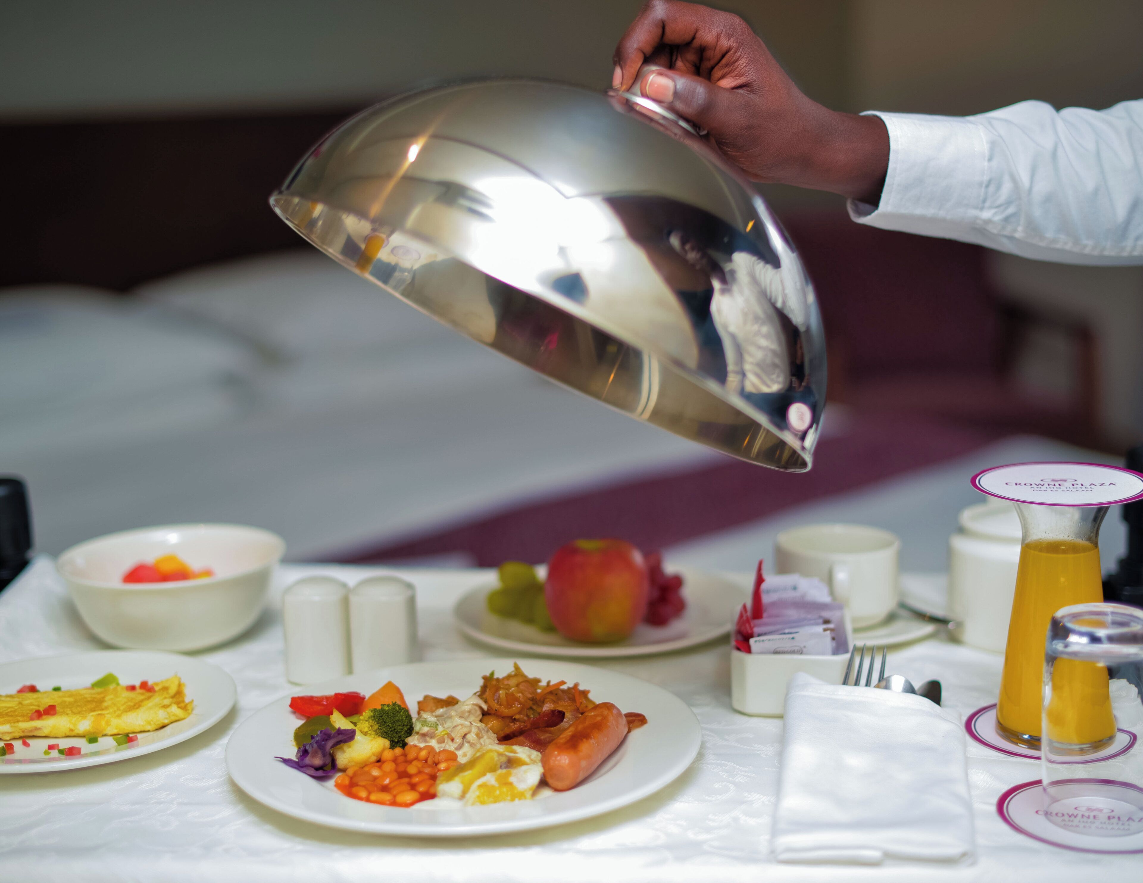 A breakfast setting with plates of food, a glass of orange juice, and a hand holding a silver bowl.