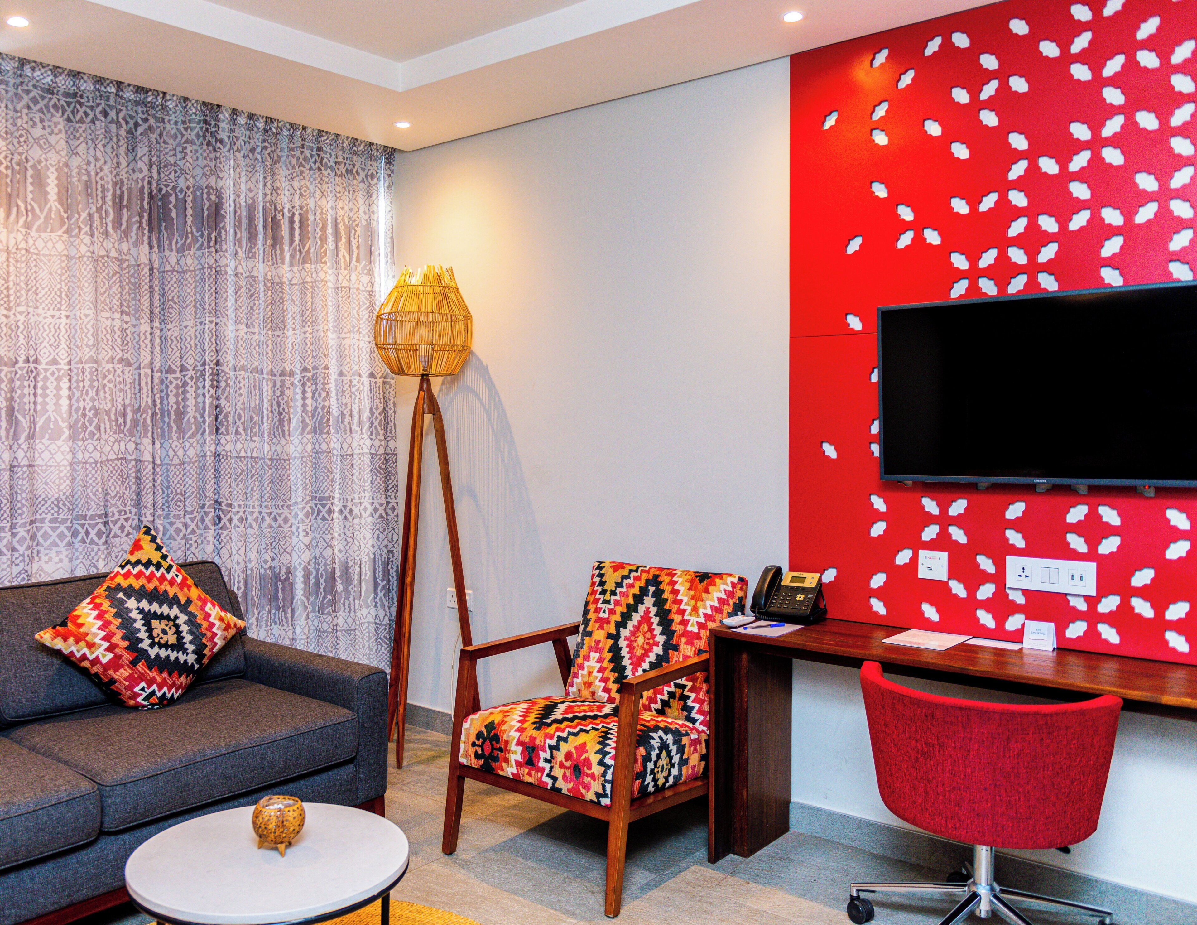 A modern living room with a sofa, a patterned armchair, a small round table, a floor lamp, and a desk with a red chair.