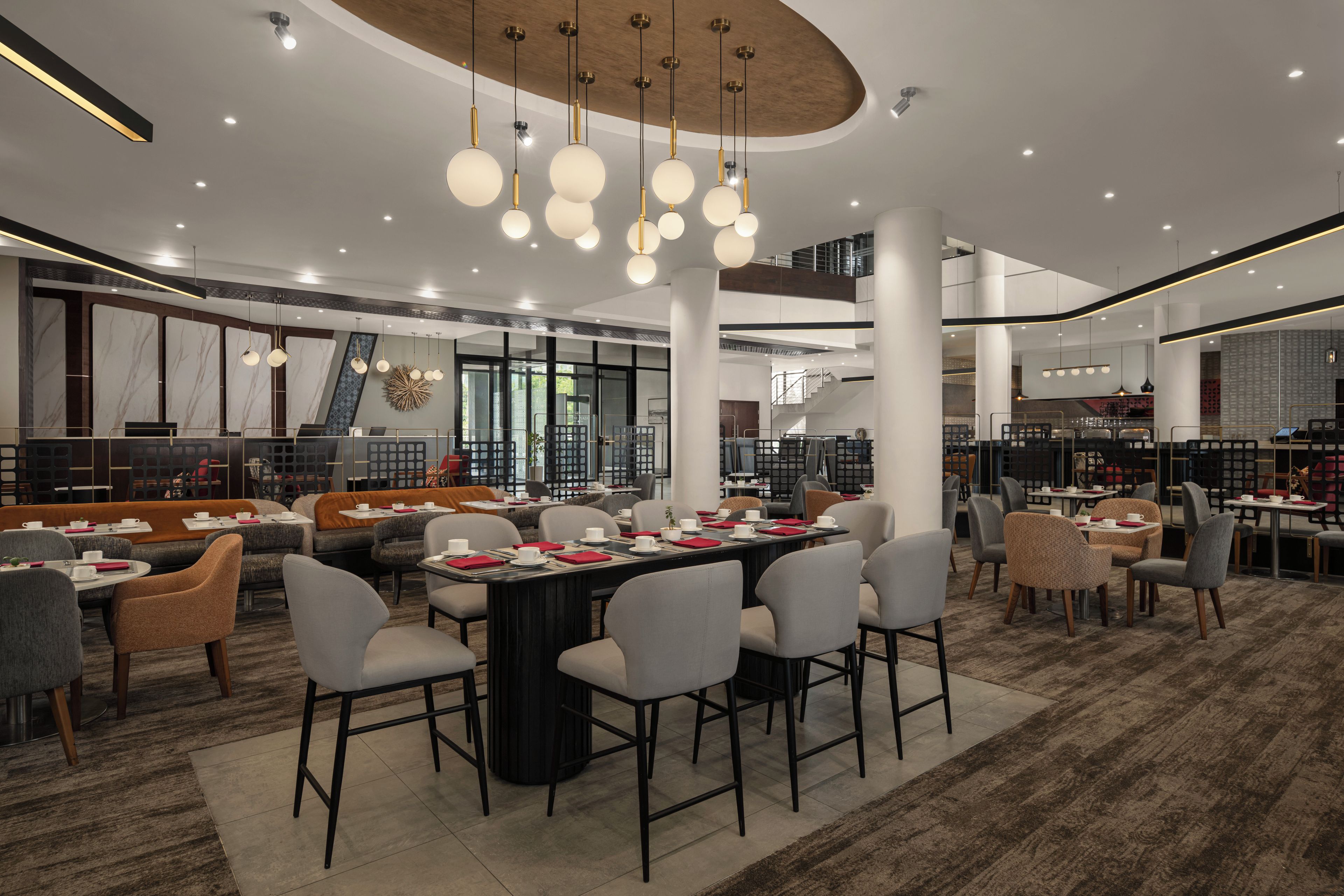 A modern hotel lobby with a central dining area, seating arrangements, and a staircase leading to an upper level.