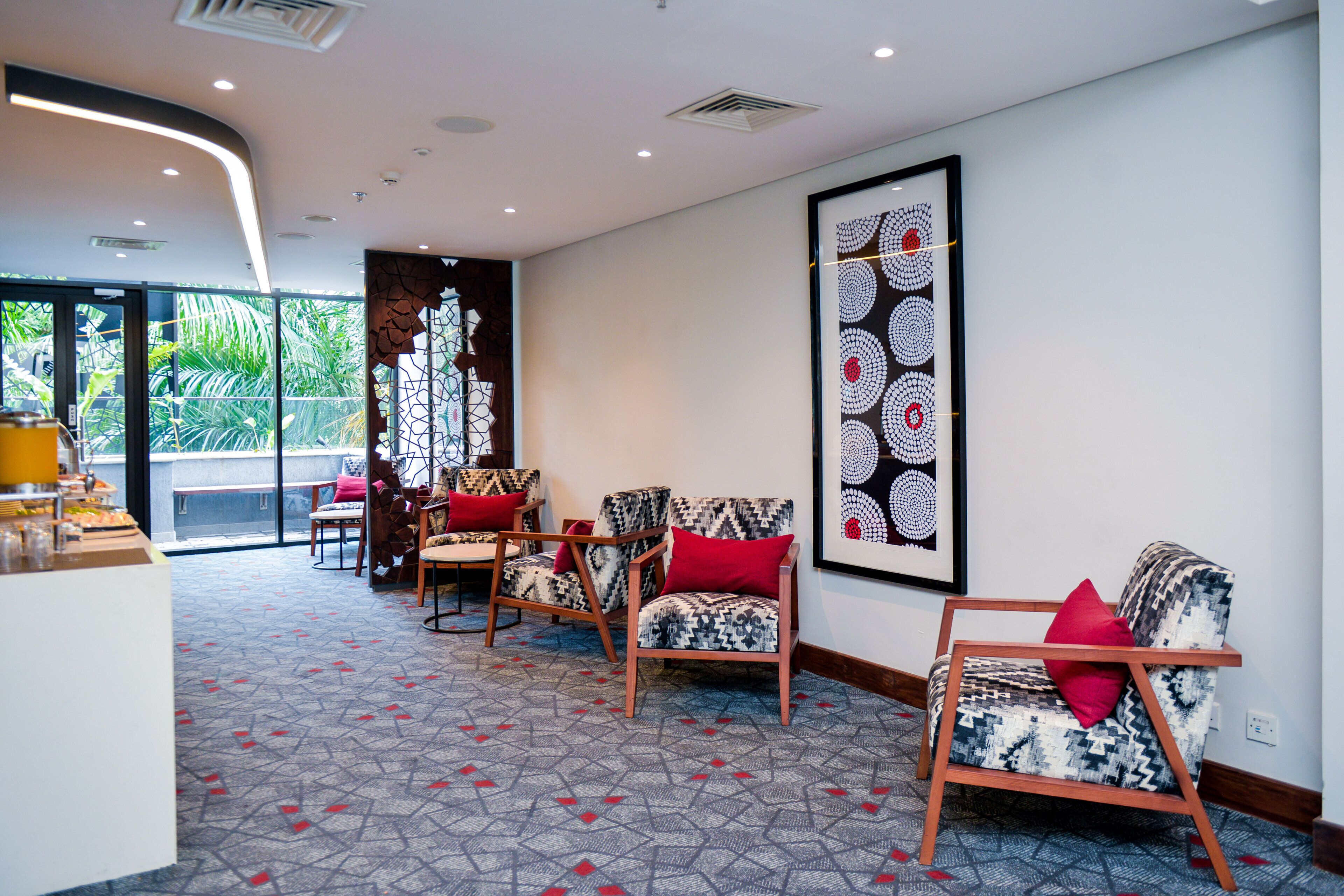 Un hall d’hôtel moderne avec un tapis à motifs, des chaises en bois dotées de coussins rouges et une grande œuvre d’art abstrait accrochée au mur.