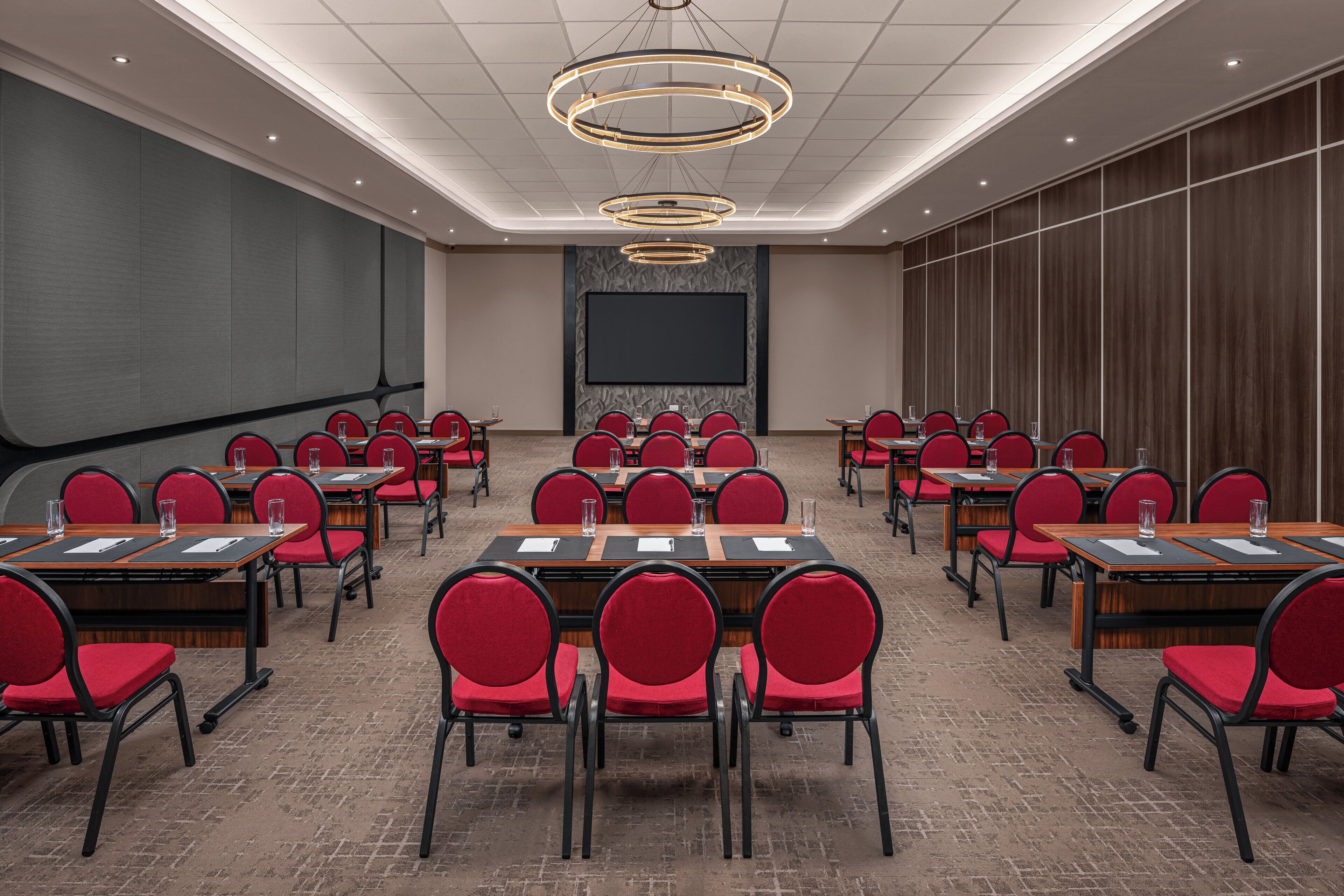 A conference room with multiple tables and chairs, a large screen, and a modern chandelier.