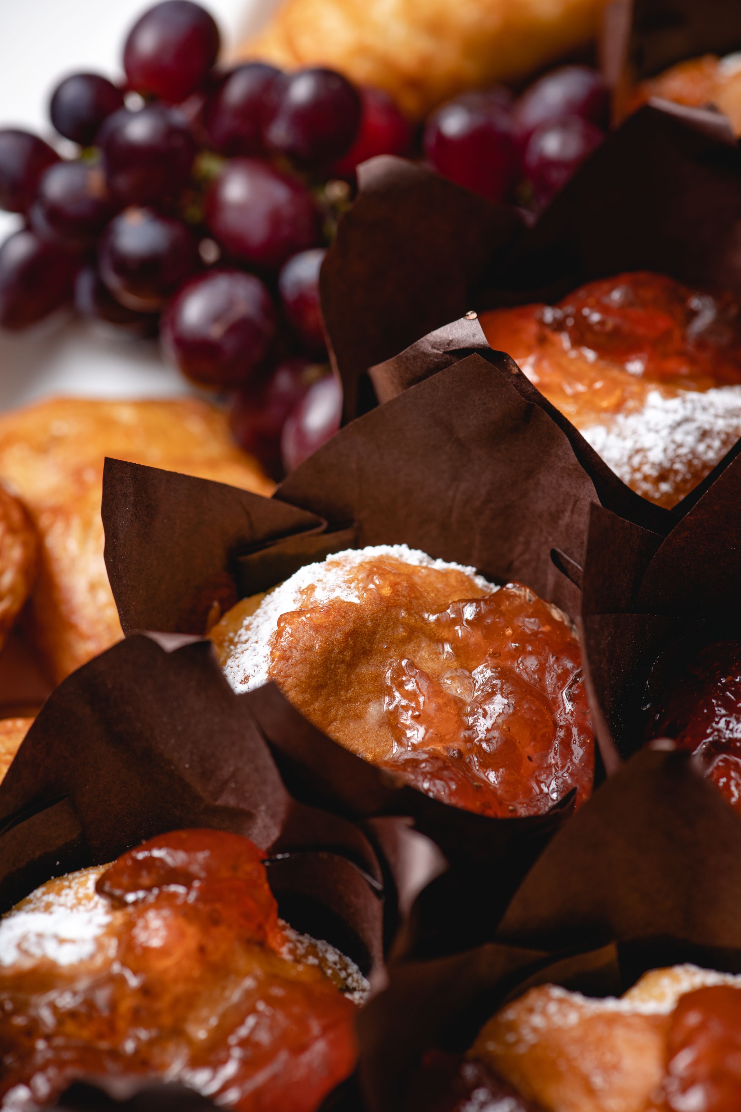 Une vue rapprochée de pâtisseries dans des sacs en papier brun, saupoudrées de sucre glace, et, à l’arrière-plan, une grappe de raisins rouges.