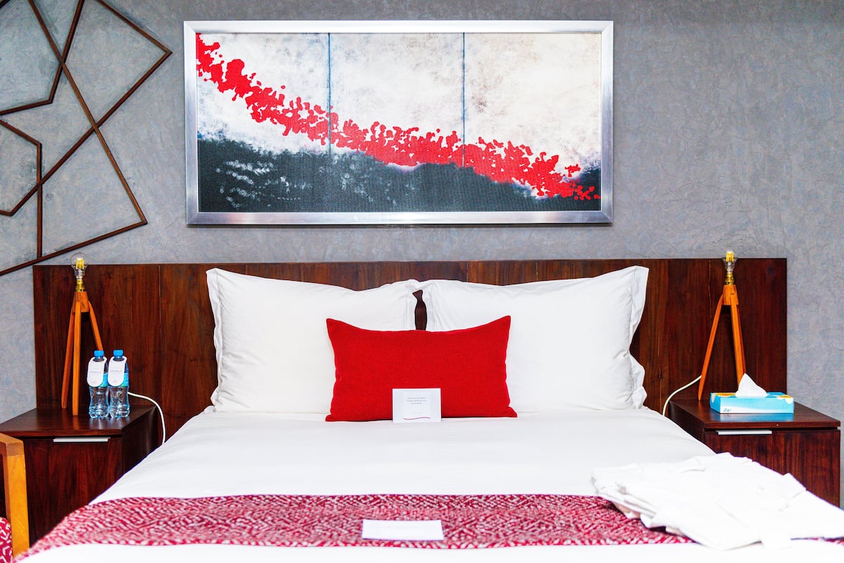 Une chambre d’hôtel avec un grand lit, un oreiller rouge, une œuvre d’art encadrée au-dessus du lit, et deux tables de chevet avec des bouteilles d’eau.