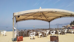 Area pranzo in spiaggia con tenda parasole, sedie e tavoli di plastica.
