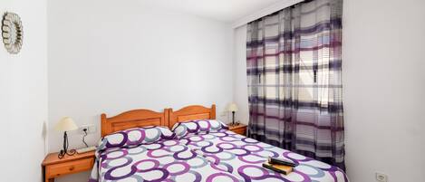 Un dormitorio con una cama, mesitas de noche de madera, una lámpara y un ventanal con cortinas.