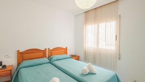 A hotel room with two beds, a wooden headboard, a nightstand with a lamp, and a large window with curtains. - Apartamentos el Rocio Casa Azahar (Alcala de Xivert)