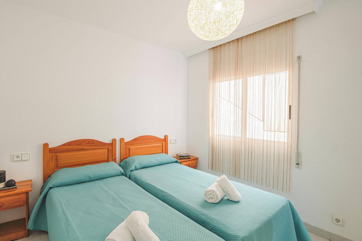 A hotel room with two beds, a wooden headboard, a nightstand with a lamp, and a large window with curtains.