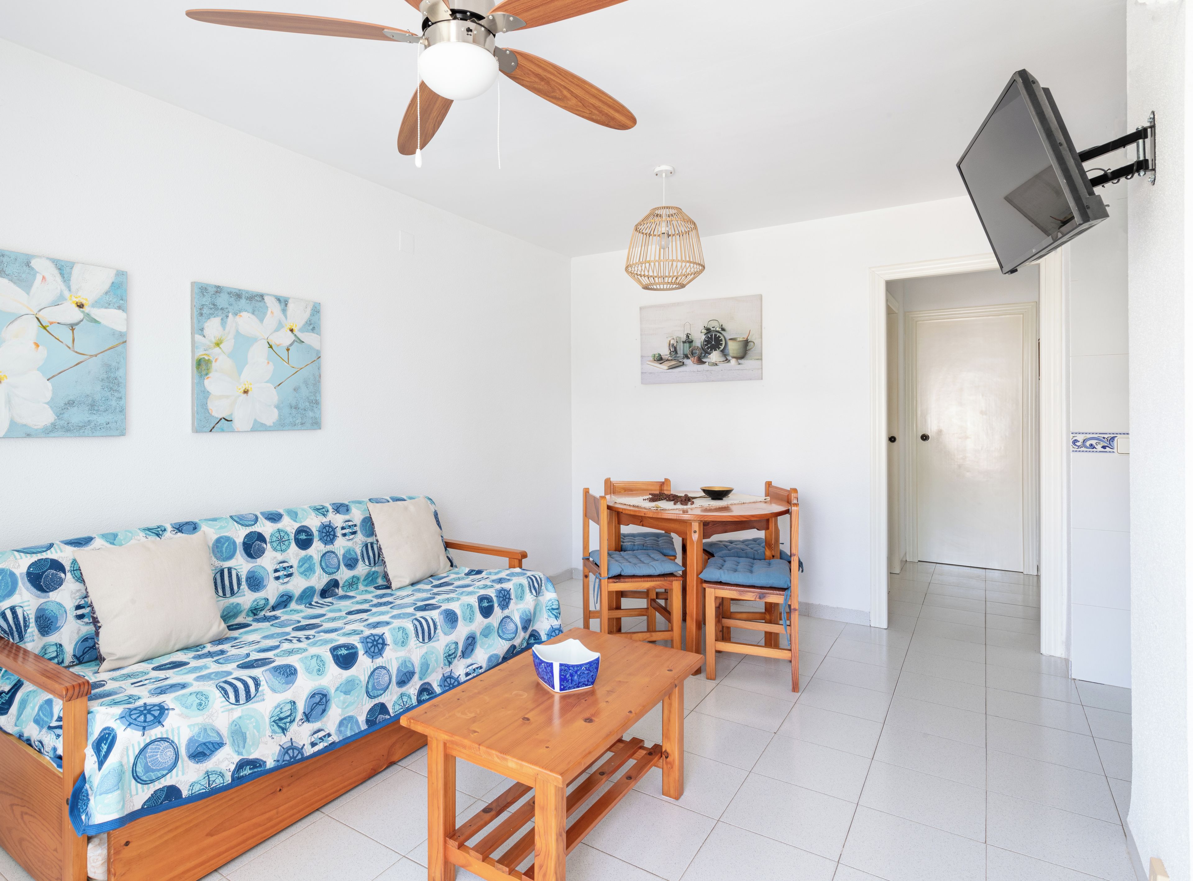 Une pièce comprenant un canapé en bois, une table en bois, un ventilateur de plafond, un téléviseur fixé au mur et deux tableaux accrochés au mur.