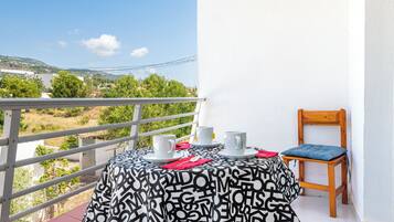 A balcony with a table set for two, a chair, and a view of a landscape with buildings and trees.