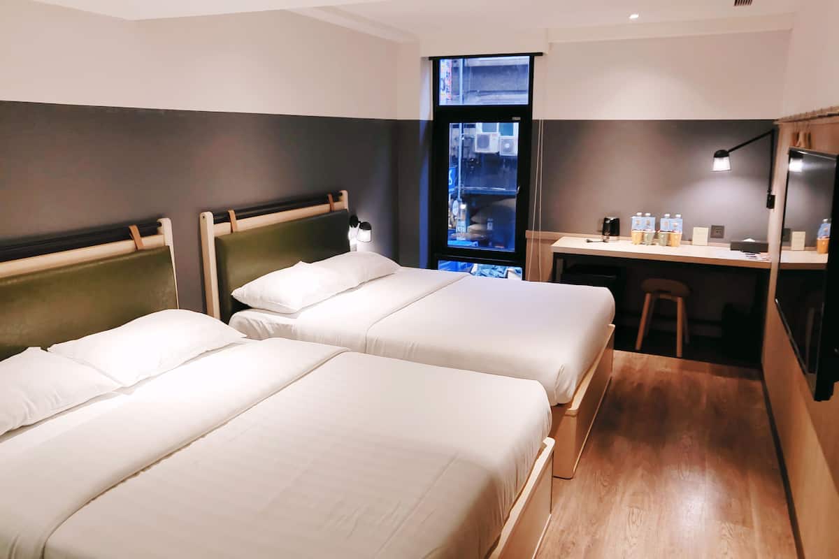 A hotel room with two beds, a desk, and a window with a view of the city.