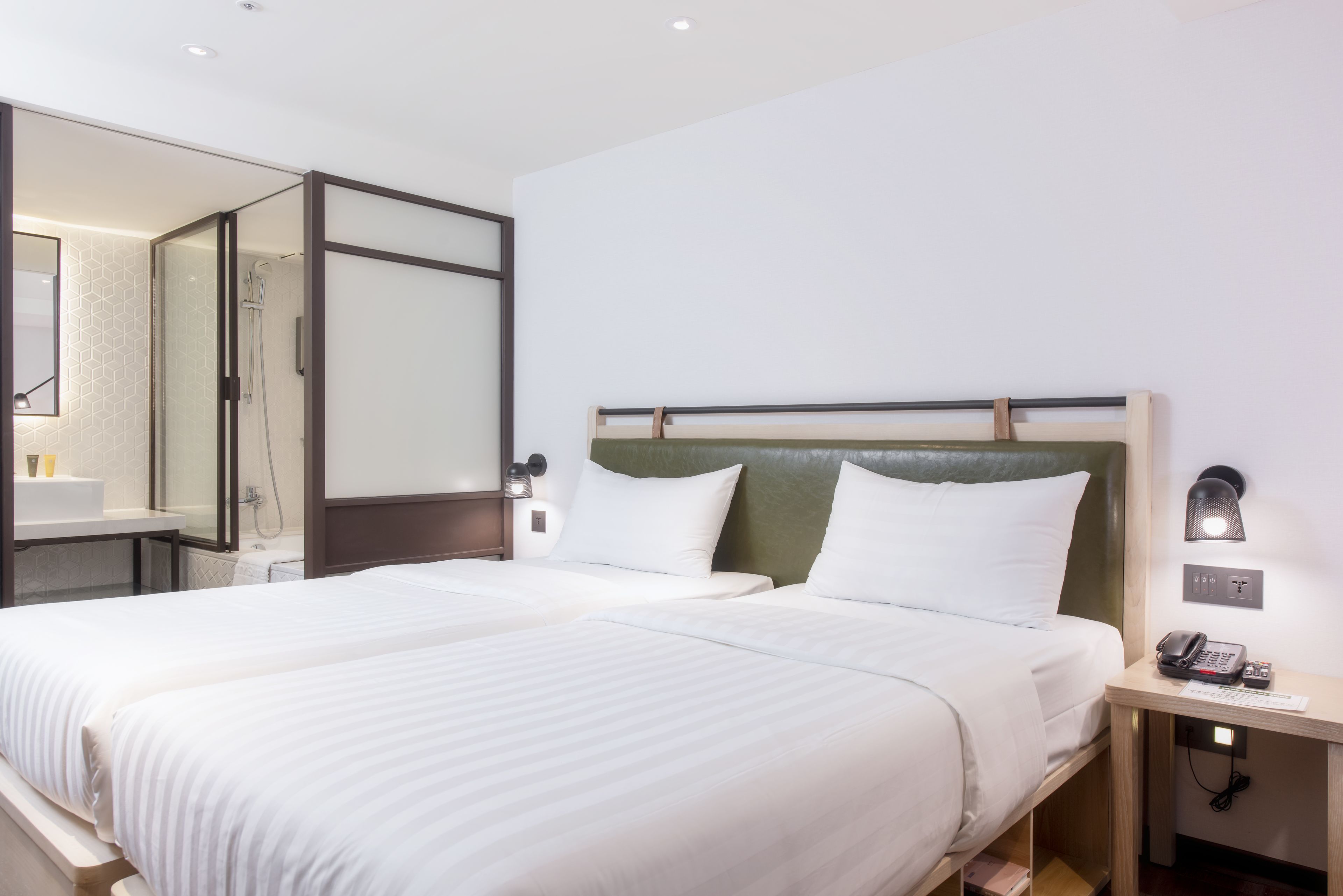 A hotel room with two beds, a shower area, a bedside table with a phone, and a wall-mounted light.