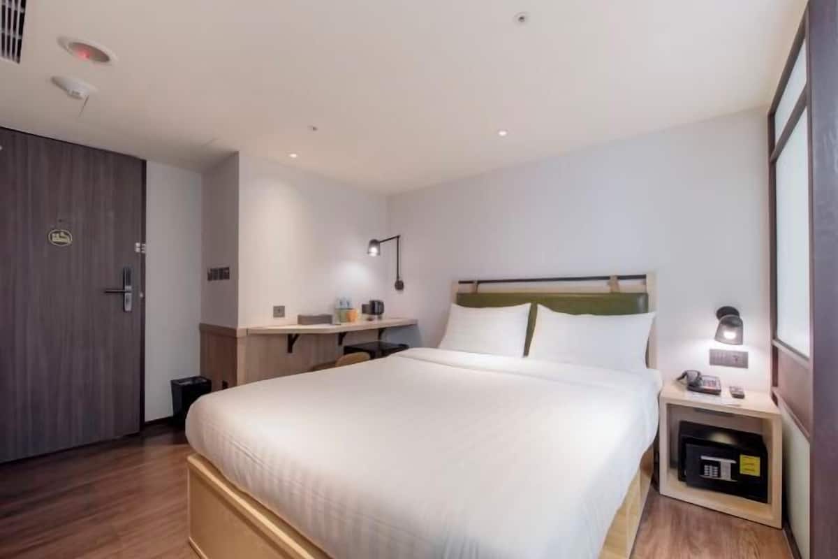 A hotel room with a large bed, a bedside table, a desk, a lamp, and a wardrobe.