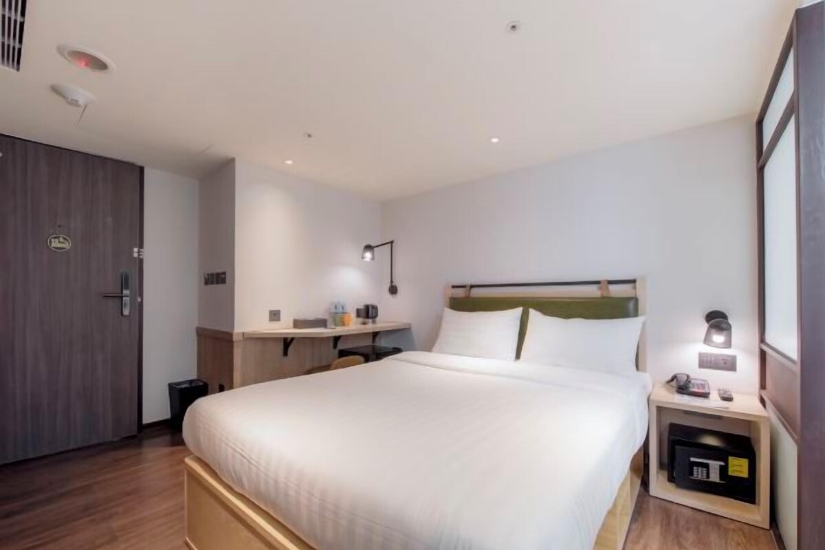 A hotel room with a large bed, a bedside table, a desk, a lamp, and a wardrobe.