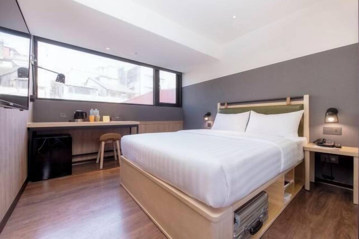 A modern hotel room with a large bed, a desk, a chair, and a view through a large window.