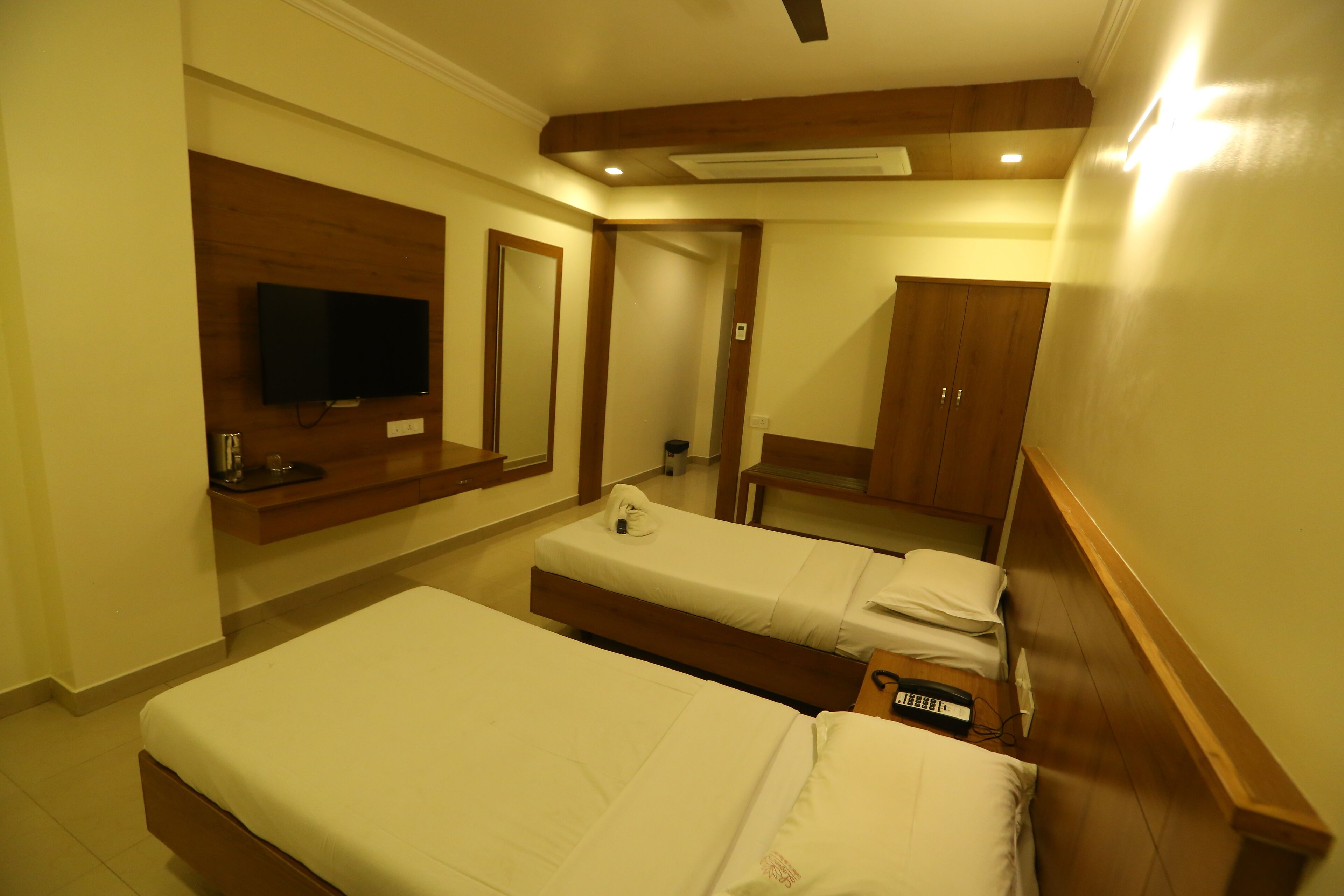A hotel room with two single beds, a TV mounted on the wall, a wooden wardrobe, and a desk with a chair.